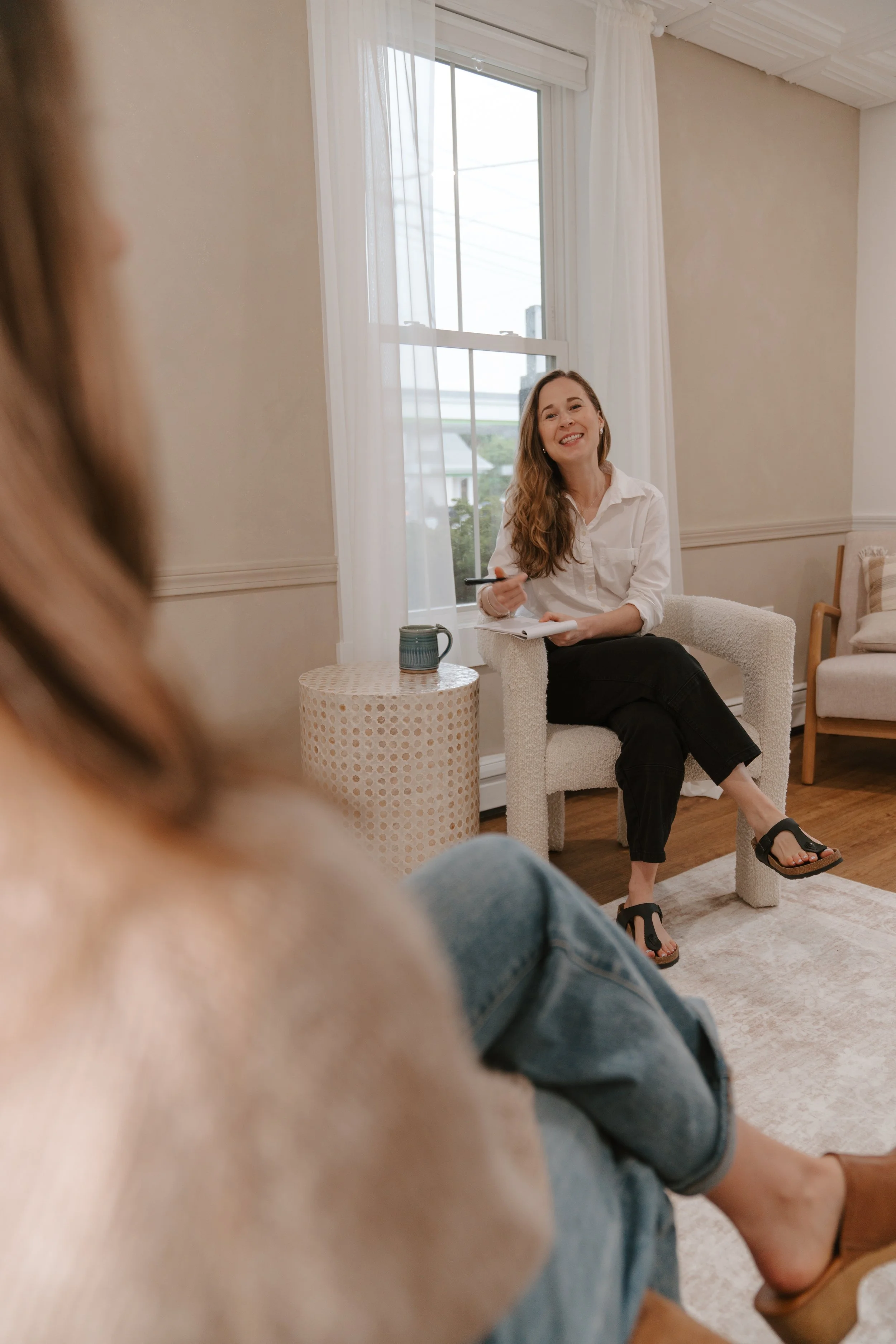 A female therapist with long brown hair smiling while sitting on a beige armchair, holding a notepad and a pen in a bright room with large window and white curtains, conducting therapy with another person partially visible in the foreground.