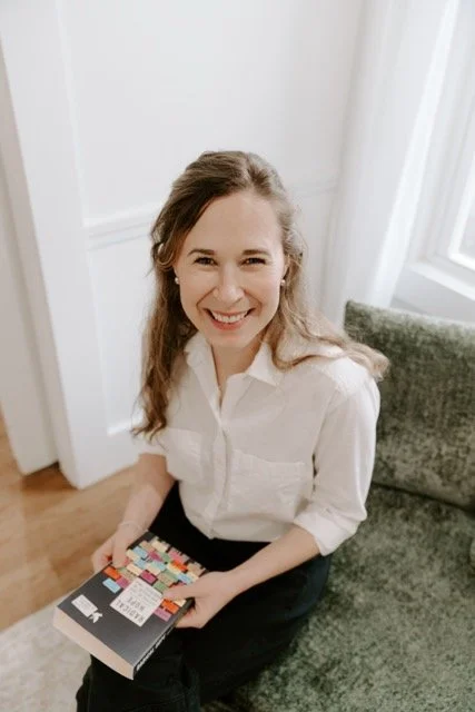 A female therapist with long, wavy brown hair, wearing a white button-up shirt, sitting on a green sofa, holding a colorful therapy book, smiling at the camera.
