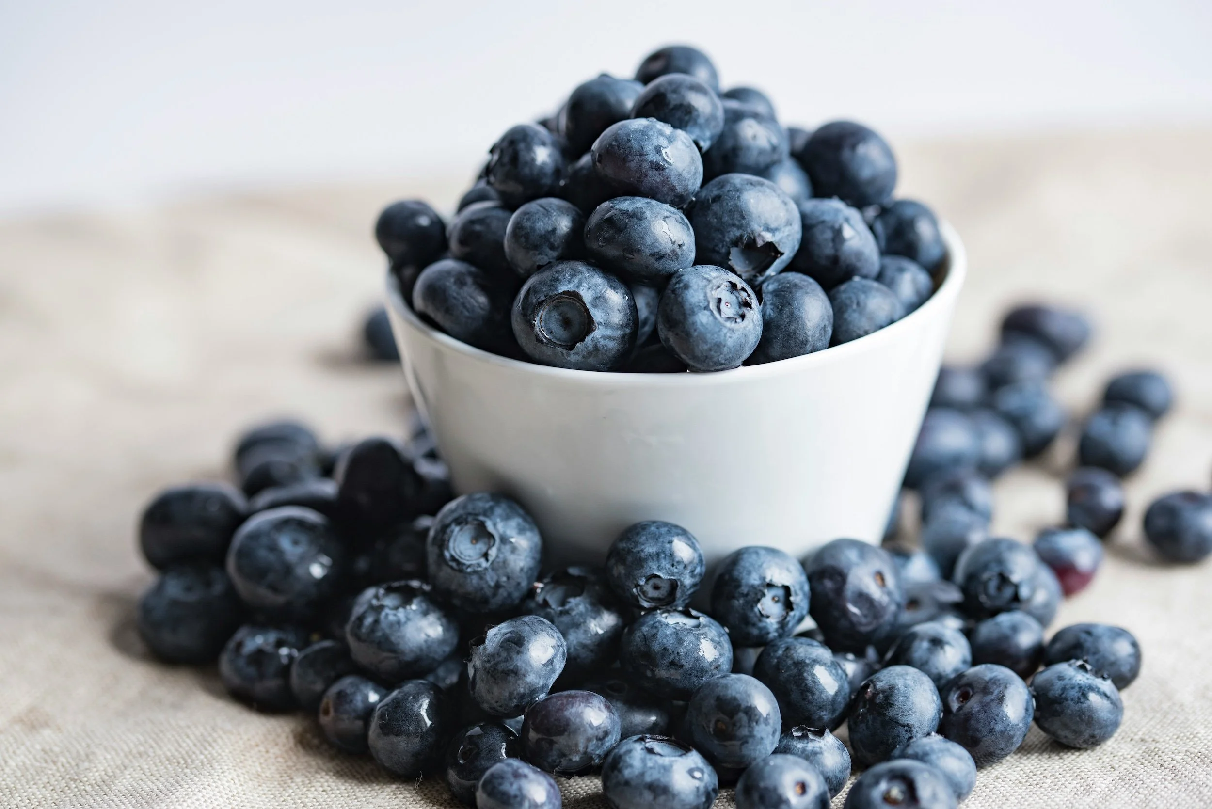A white bowl filled with blueberries, with more blueberries scattered on a beige surface.