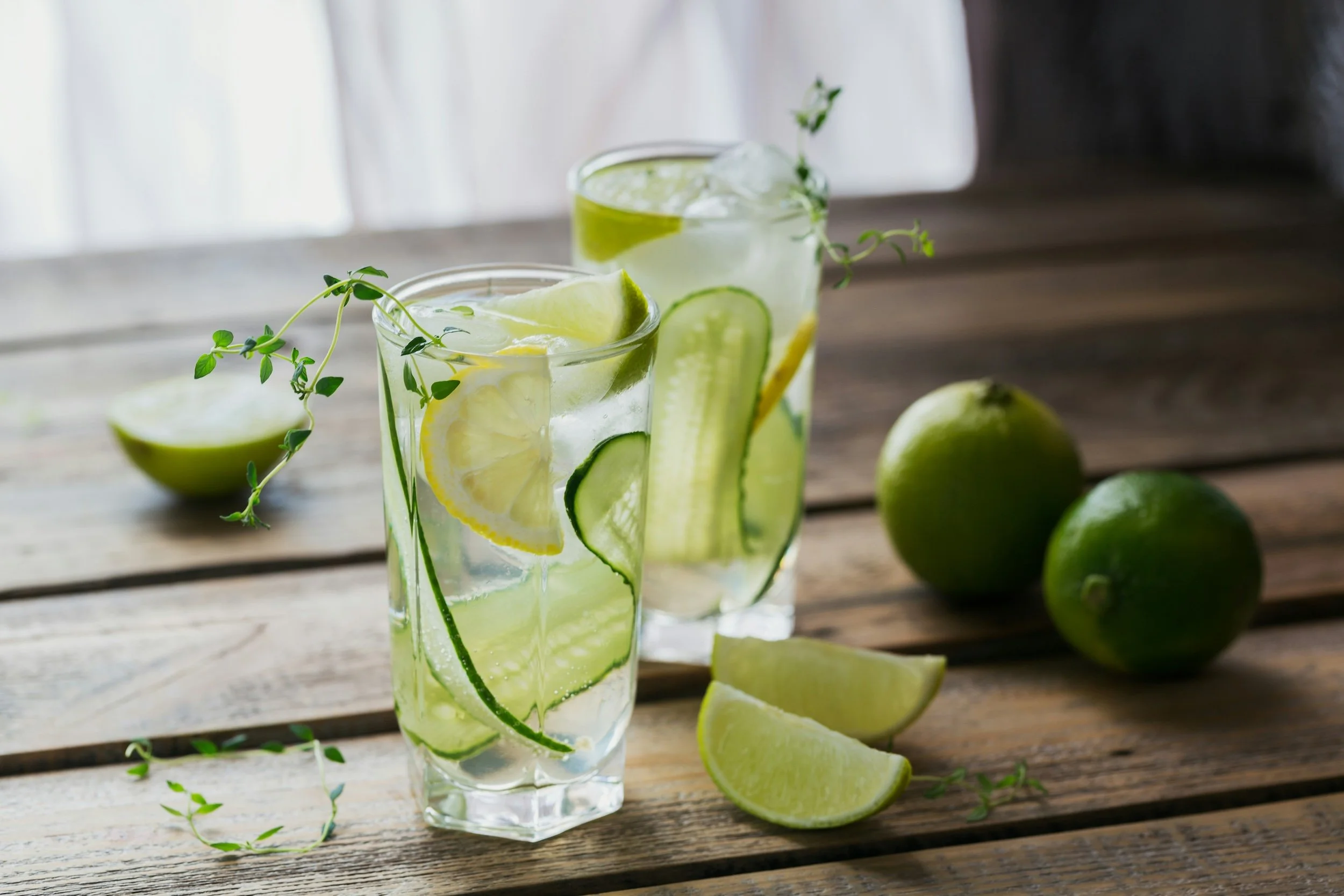 Two glasses of cucumber and lemon infused water garnished with thyme on a rustic wooden table, with fresh limes and lime wedges nearby.