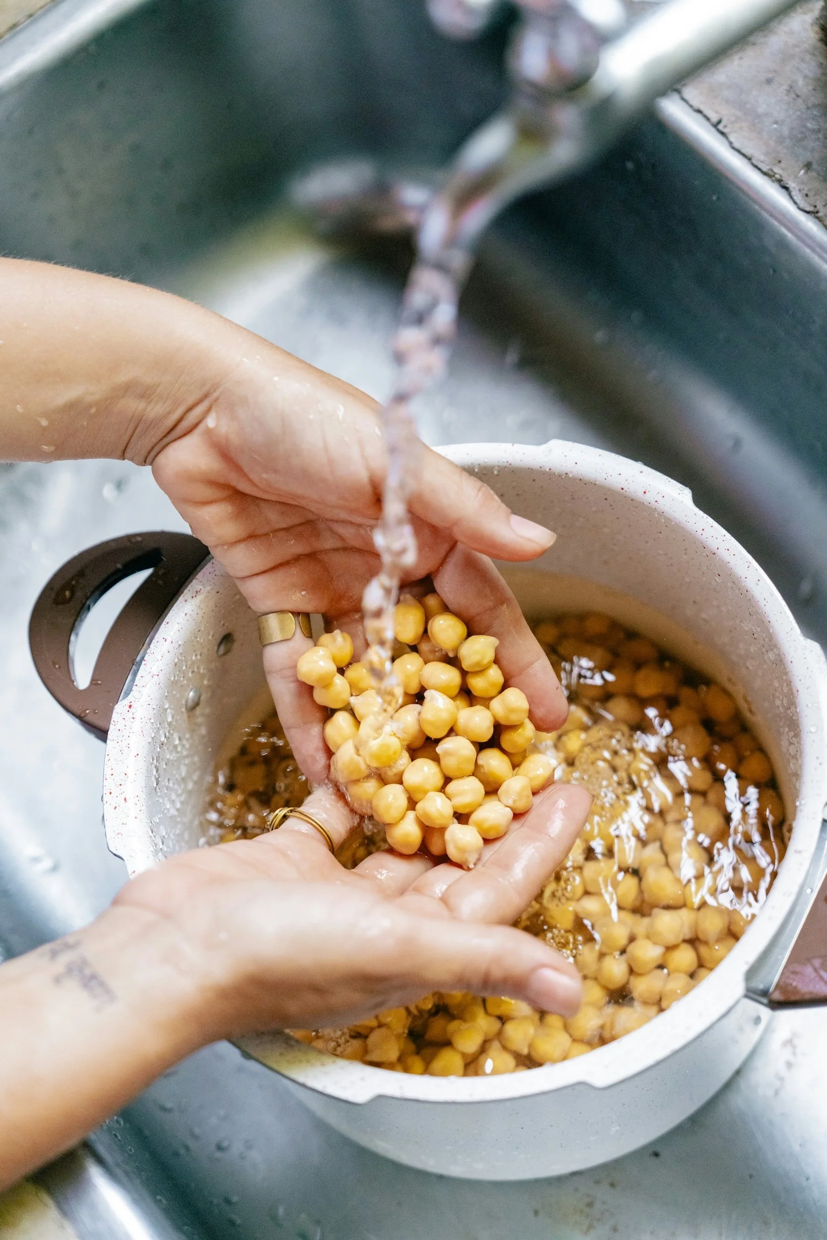 Person rinsing chickpeas in a white colander under a kitchen faucet.