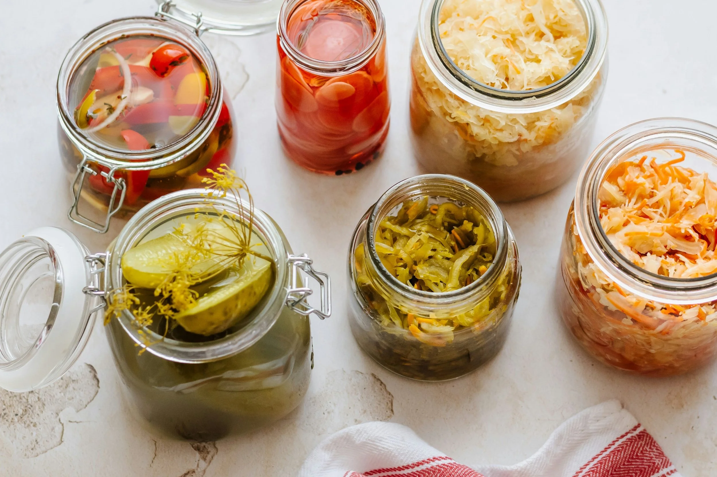 Various jars of homemade pickles, sauerkraut, and fermented vegetables on a white surface with a red and white cloth.