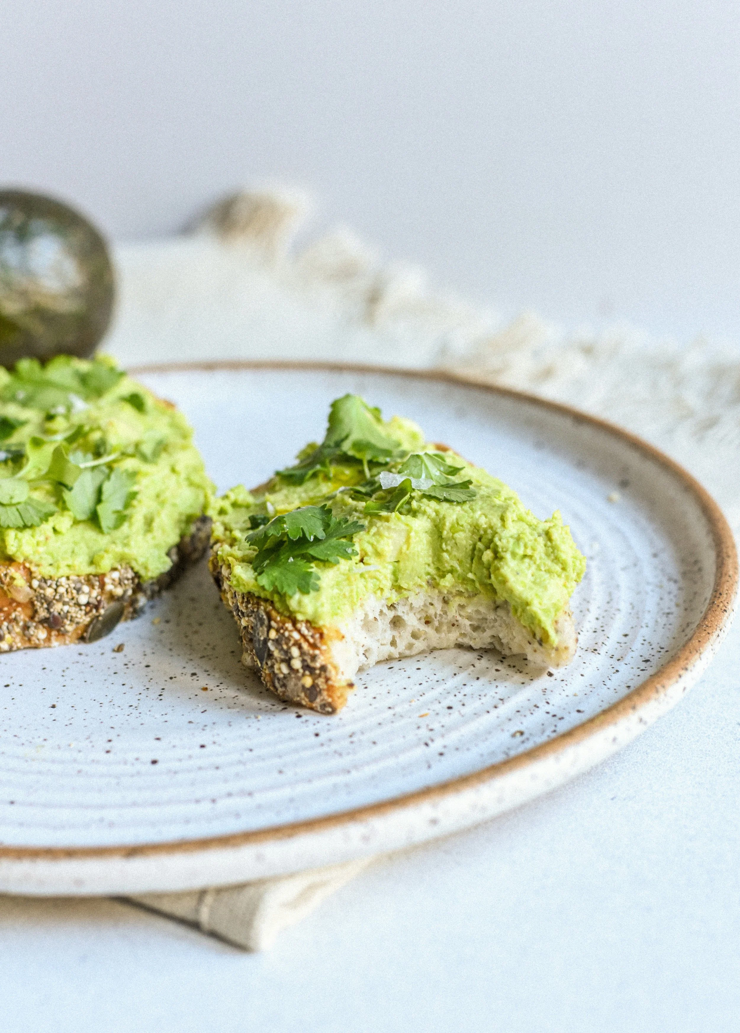 Half-eaten slice of avocado toast with cilantro and seed-crusted bread on a ceramic plate.