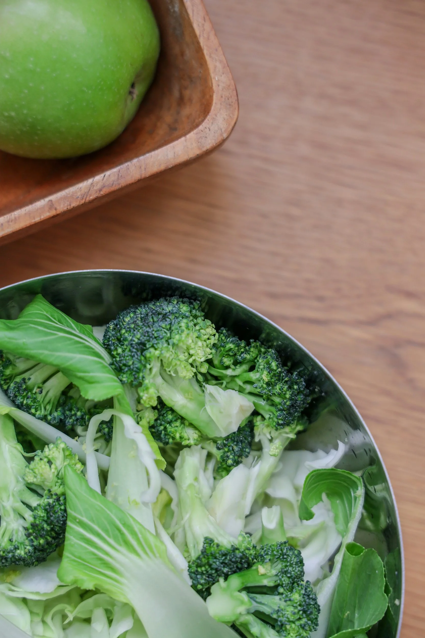 A bowl of chopped broccoli and cabbage on a wooden surface, with a green apple in a wooden bowl nearby.