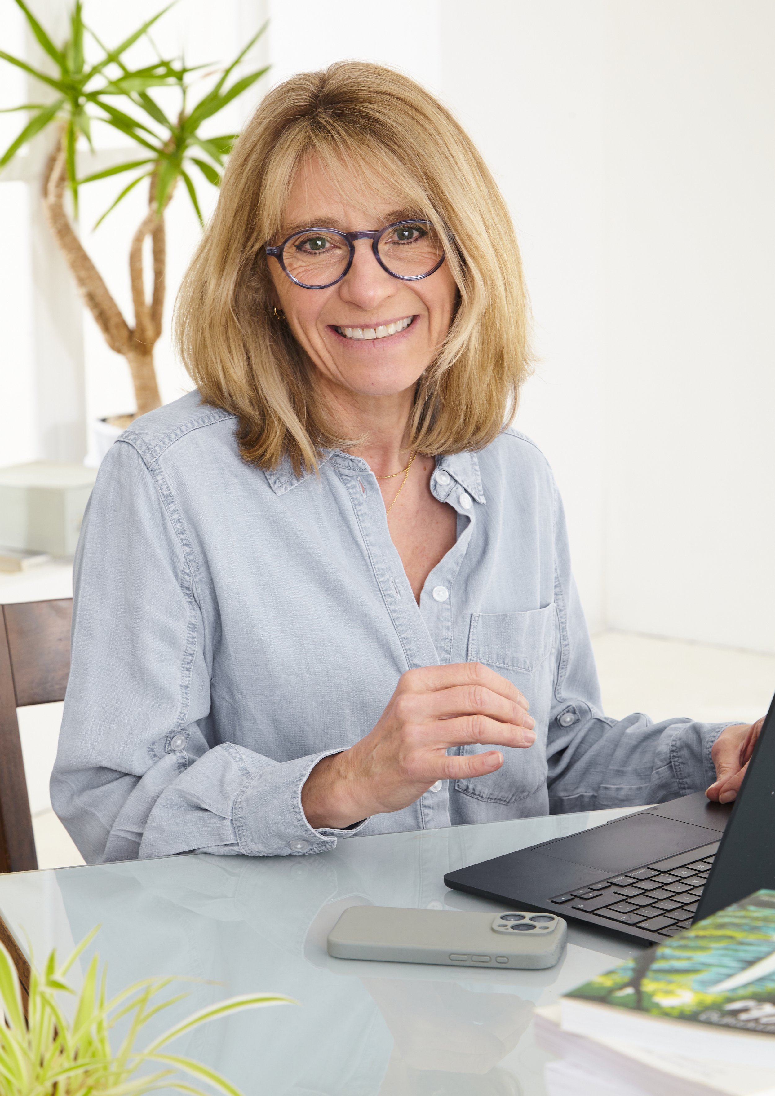 A woman with glasses and shoulder-length blonde hair smiling while sitting at a desk with a laptop, a smartphone, and a book.