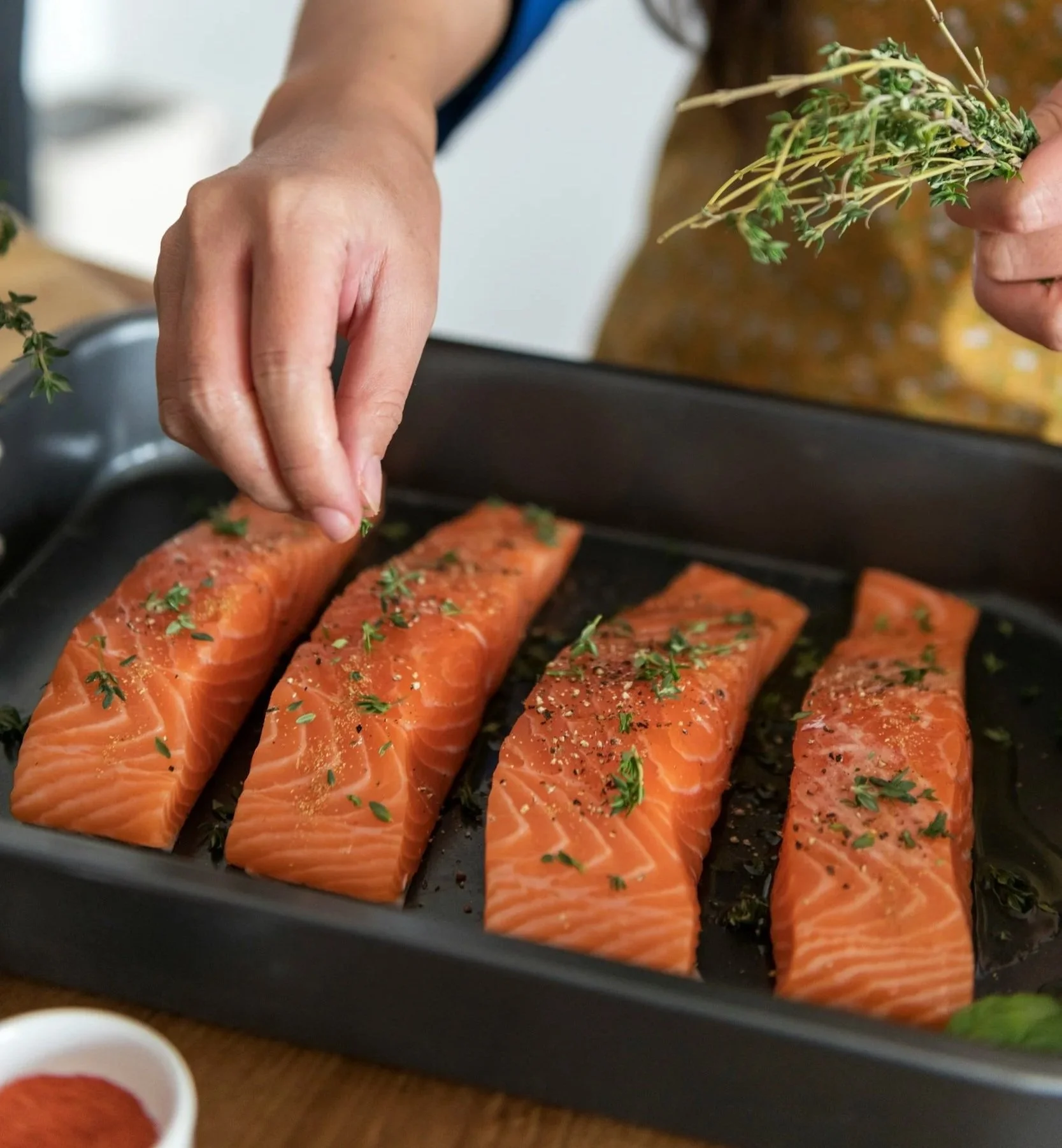 Person seasoning raw salmon fillets with herbs and spices on a baking sheet.