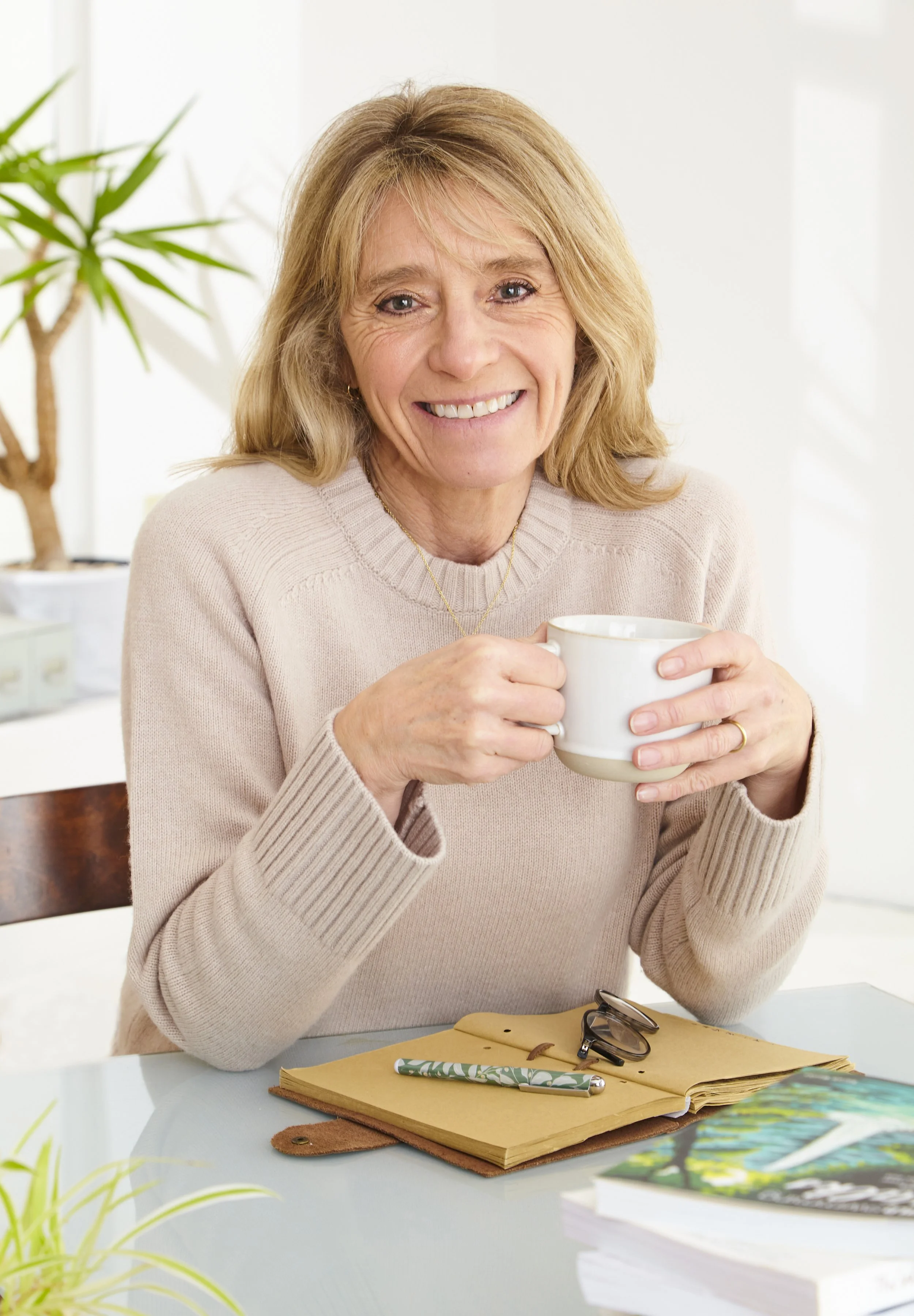 An older woman with blonde hair smiling and holding a white mug in a bright, cozy indoor setting. There are glasses, a pen, an open notebook, and books on the table in front of her.