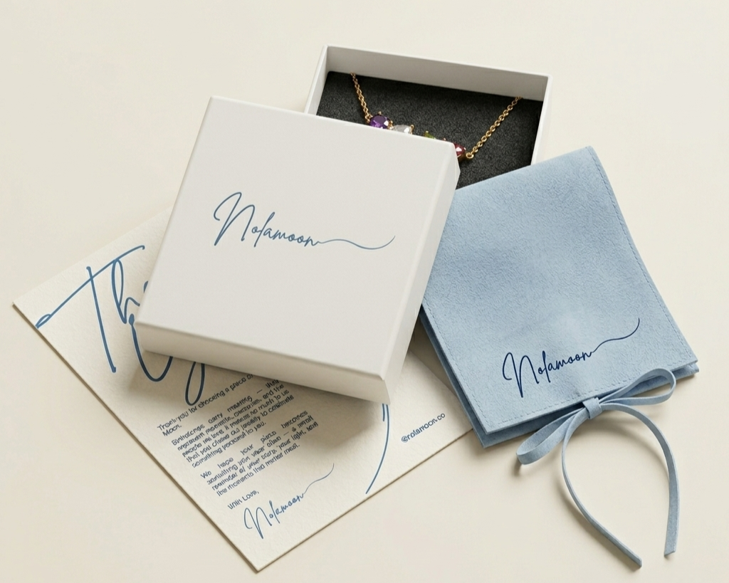 Jewelry box with a necklace inside, a blue cloth with a signature, and an informational card, all branded with 'Nadamon'