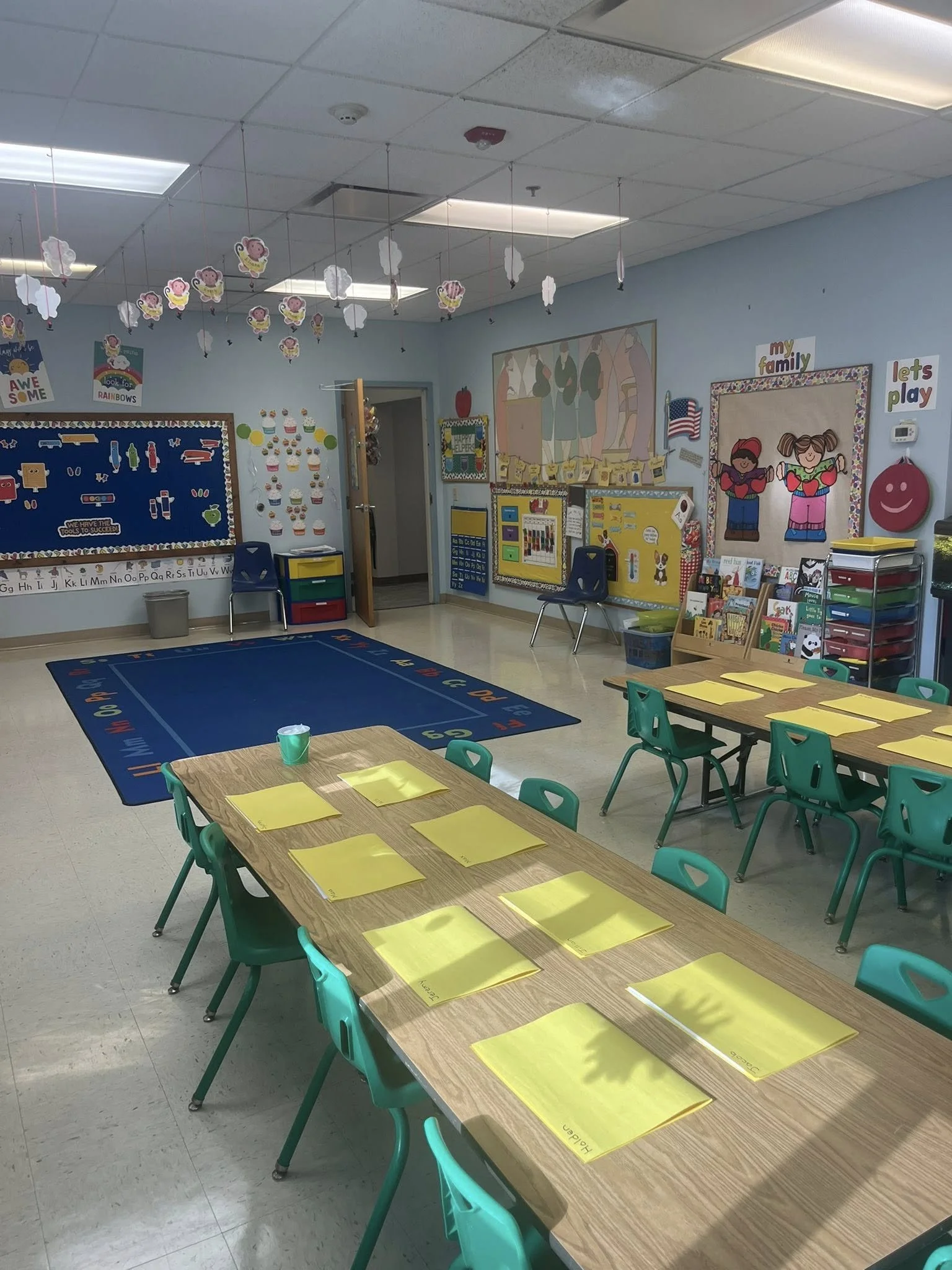 Colorful kindergarten classroom with tables set for activities, a blue rug, decorative ceiling hanging animals, educational posters, and colorful storage bins.