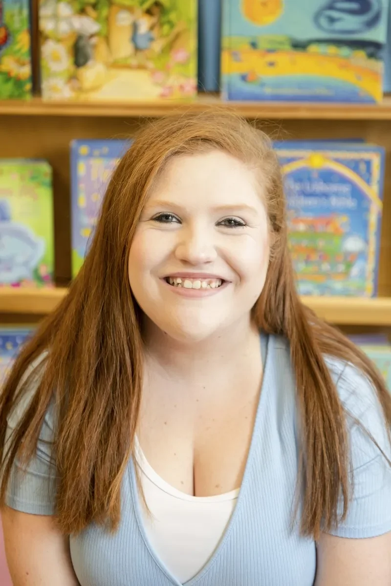A young woman with long red hair smiling in front of a bookshelf filled with colorful children's books.