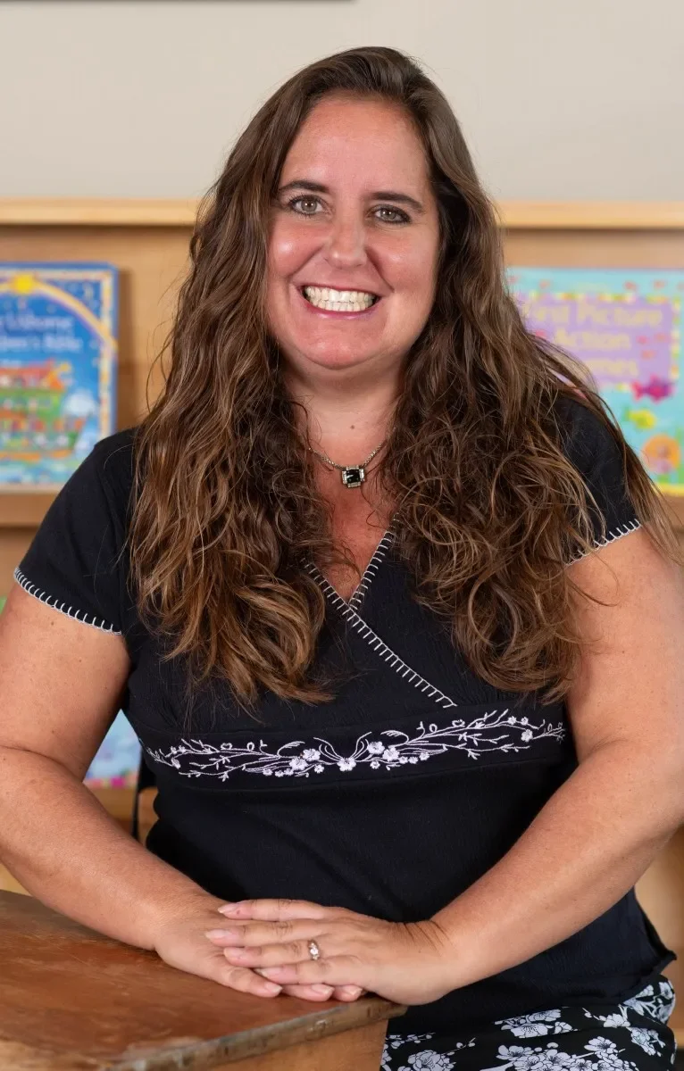 A woman with long, curly brown hair smiling and sitting at a wooden table with her hands resting on it. She is wearing a black top with white embroidery and a silver necklace. In the background, colorful children's books are visible on wooden shelves.