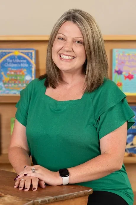 Woman with shoulder-length blonde hair smiling, wearing a green top, sitting at a wooden table with bookshelves in the background.