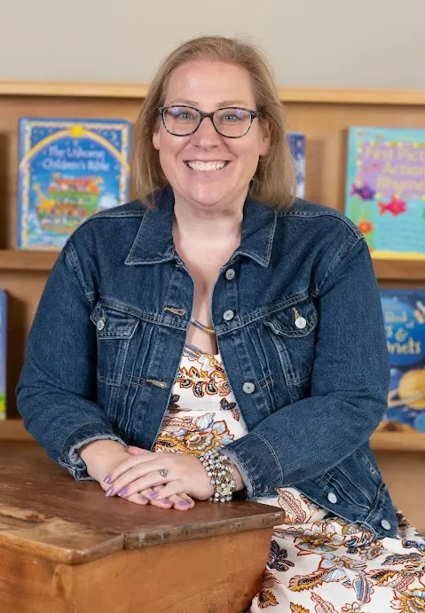 A woman with glasses and light brown hair smiling, sitting at a wooden table with bookshelves in the background.