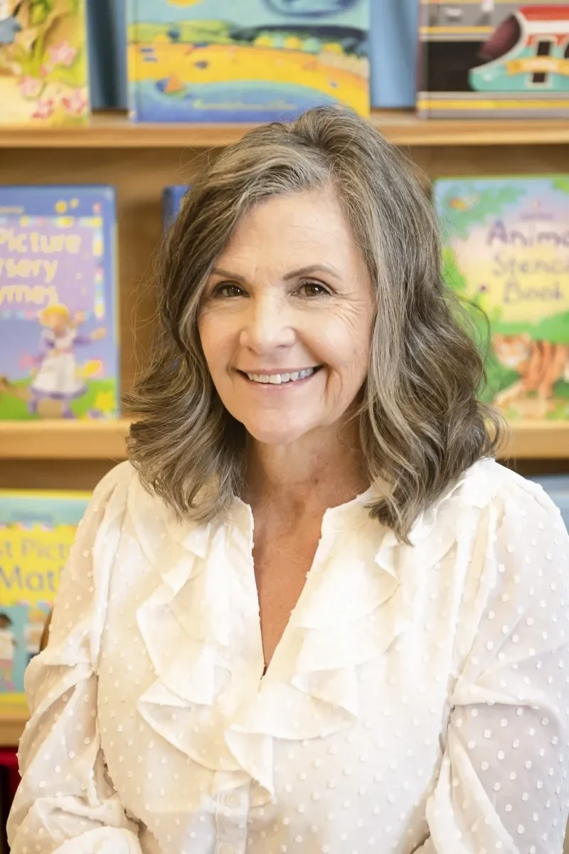 A smiling middle-aged woman with wavy gray hair, wearing a white blouse with ruffles, sitting in front of a bookshelf filled with children's books.