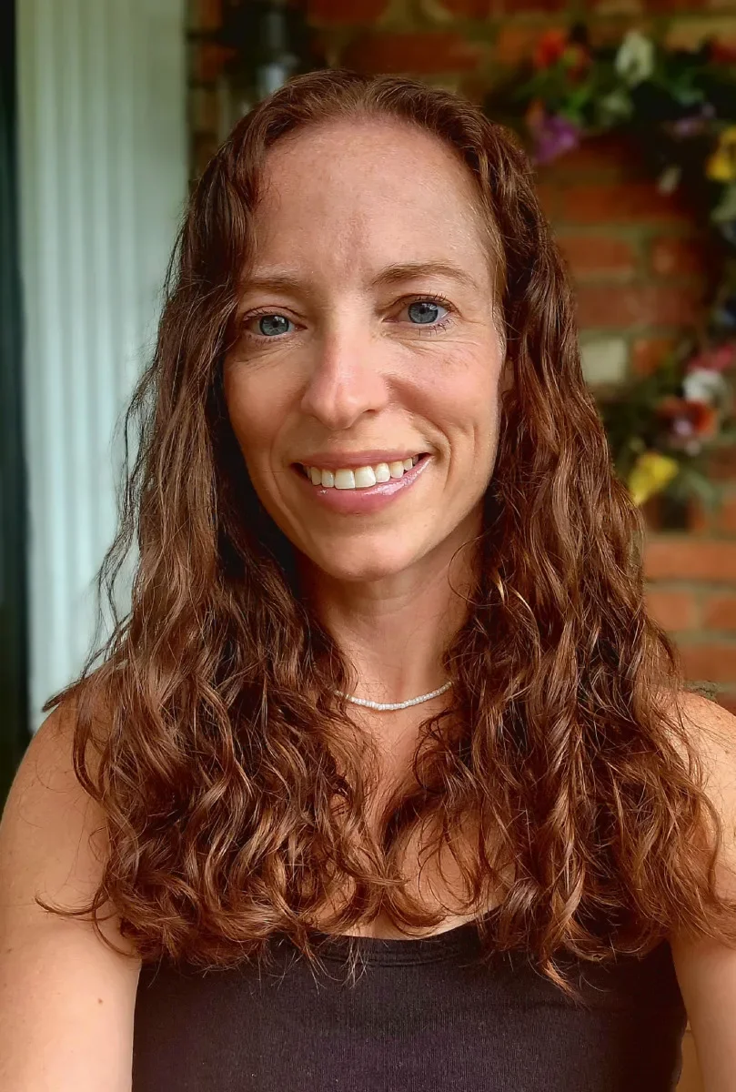 A woman with long, curly brown hair smiling, wearing a black top and a pearl necklace, standing in front of a brick wall with colorful flowers.