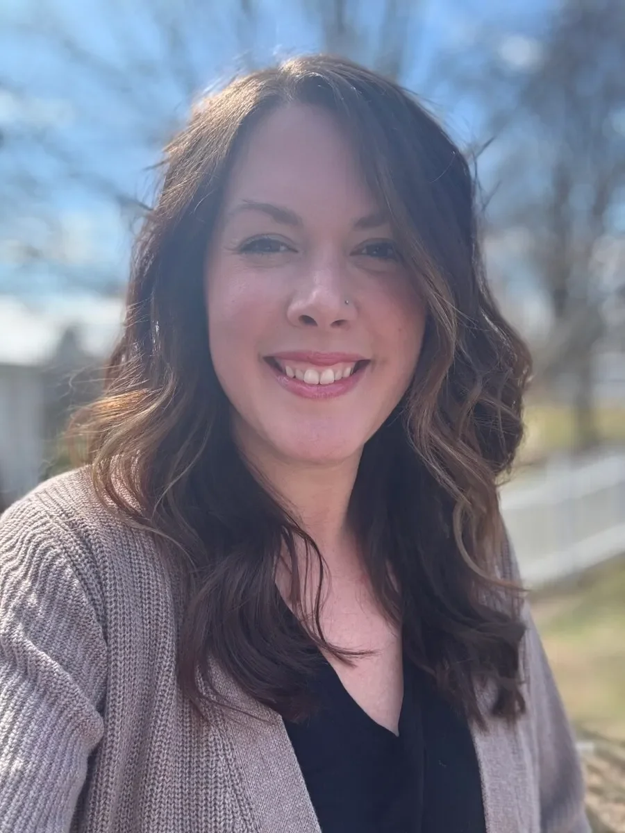 A woman with shoulder-length wavy brown hair, smiling outdoors on a sunny day, wearing a black top and beige cardigan.