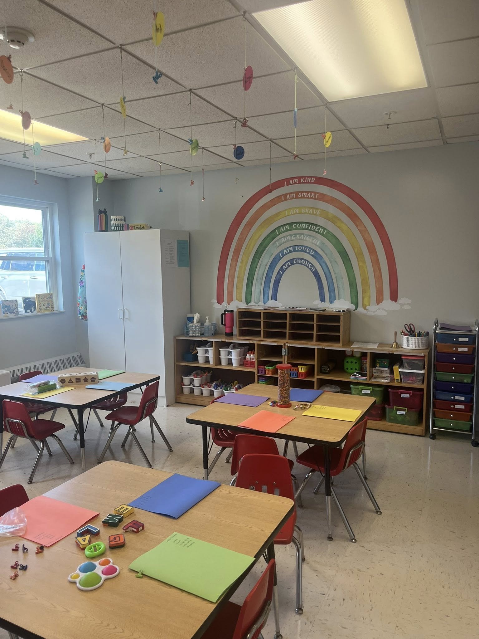 A colorful and cheerful classroom with a rainbow mural on the wall displaying positive affirmations. Several small tables with red chairs are arranged around the room, each with colored folders and educational materials. Shelves with supplies and a window letting in natural light complete the scene.