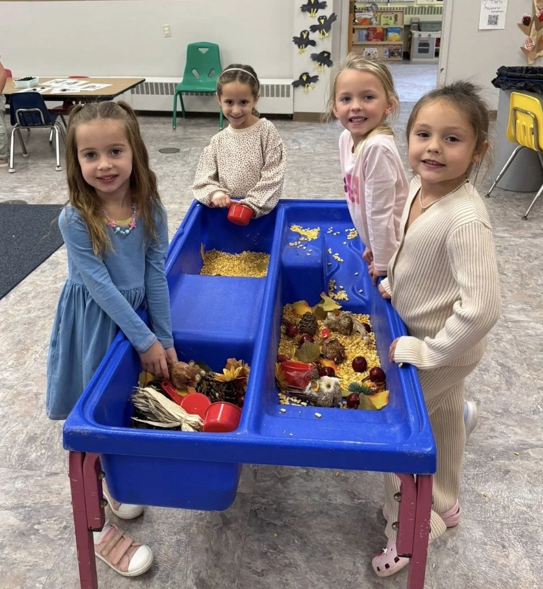 Five kids standing around a blue sensory table filled with fall-themed objects like leaves, acorns, pinecones, and artificial fruits inside a classroom.