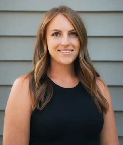 A woman with shoulder-length brown hair, wearing a black sleeveless top, standing outdoors in front of horizontal gray siding.