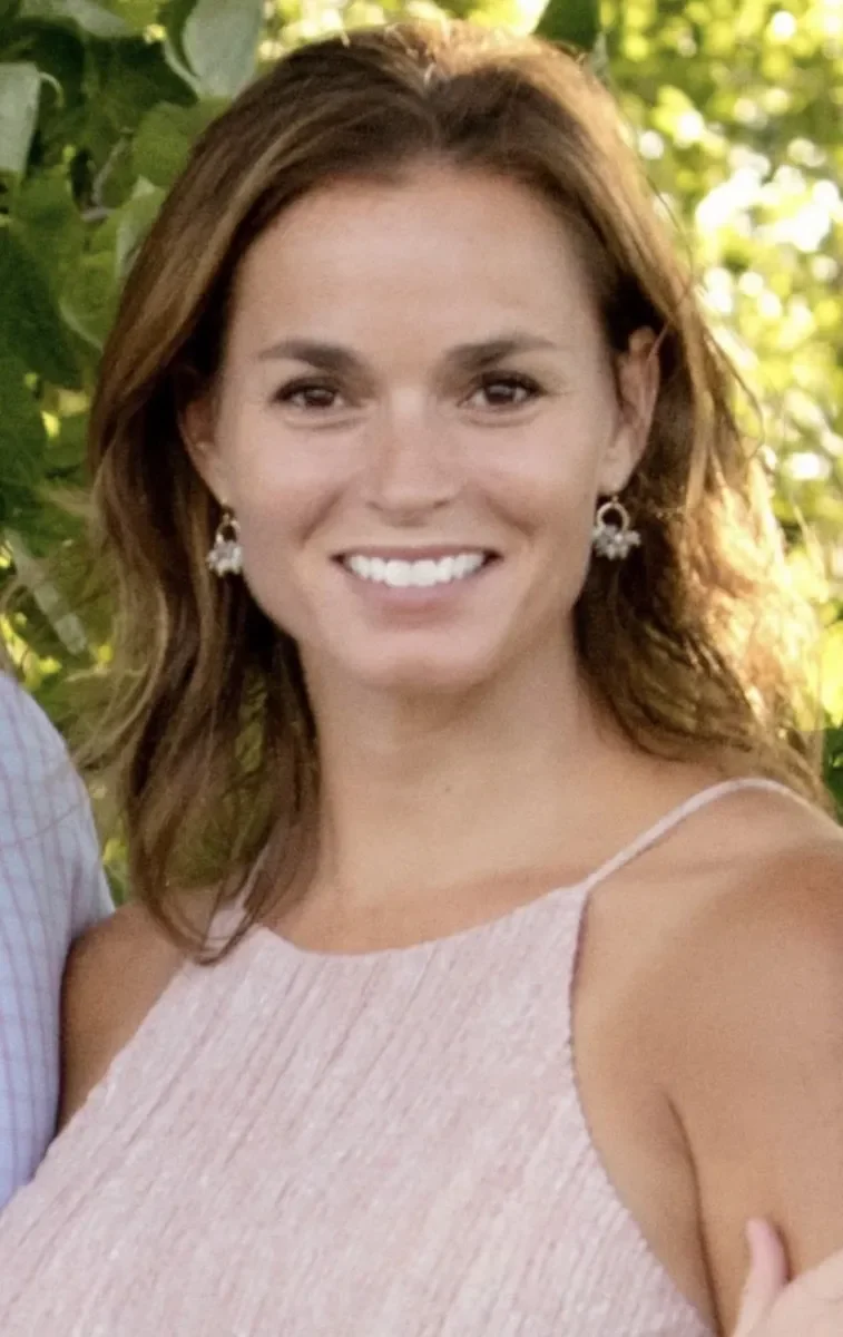 A woman with brown, wavy hair smiling outdoors, wearing a light pink sleeveless top and floral earrings, with green foliage in the background.