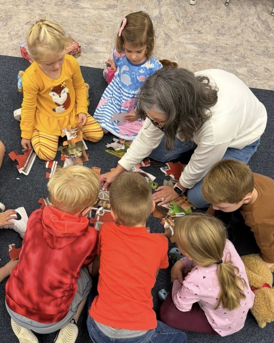A teacher and six children are sitting on a dark carpeted floor, working on a puzzle together. The teacher is leaning forward, guiding the children.