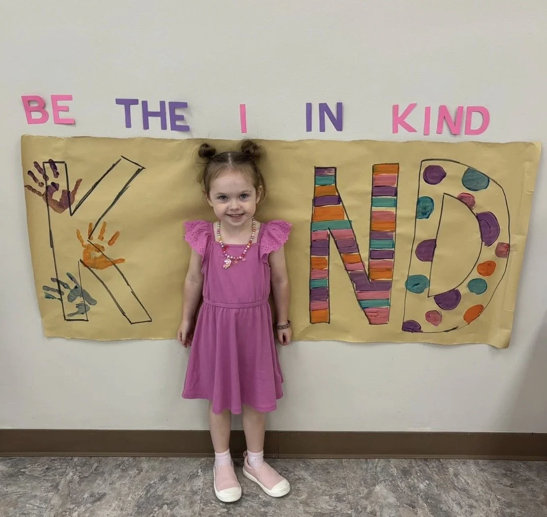 Young girl in a pink dress standing in front of a decorated wall with colorful letters spelling out 'BE THE KIND IN KIND' and large, colorful signs with decorated letters.