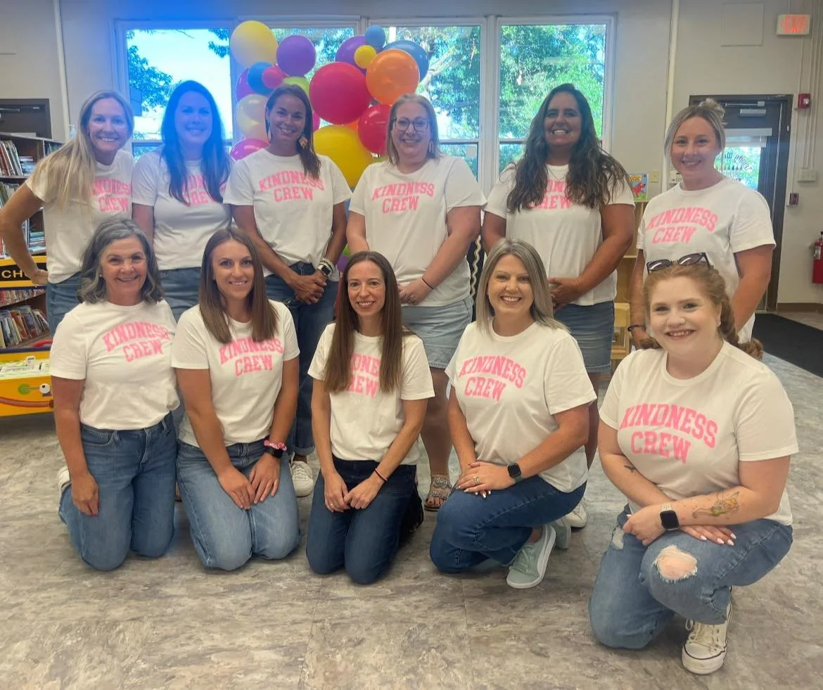 Group of ten teachers wearing matching white T-shirts with pink text that reads 'KINDNESS CREW' standing in front of colorful balloons inside a room with bookshelves and windows.