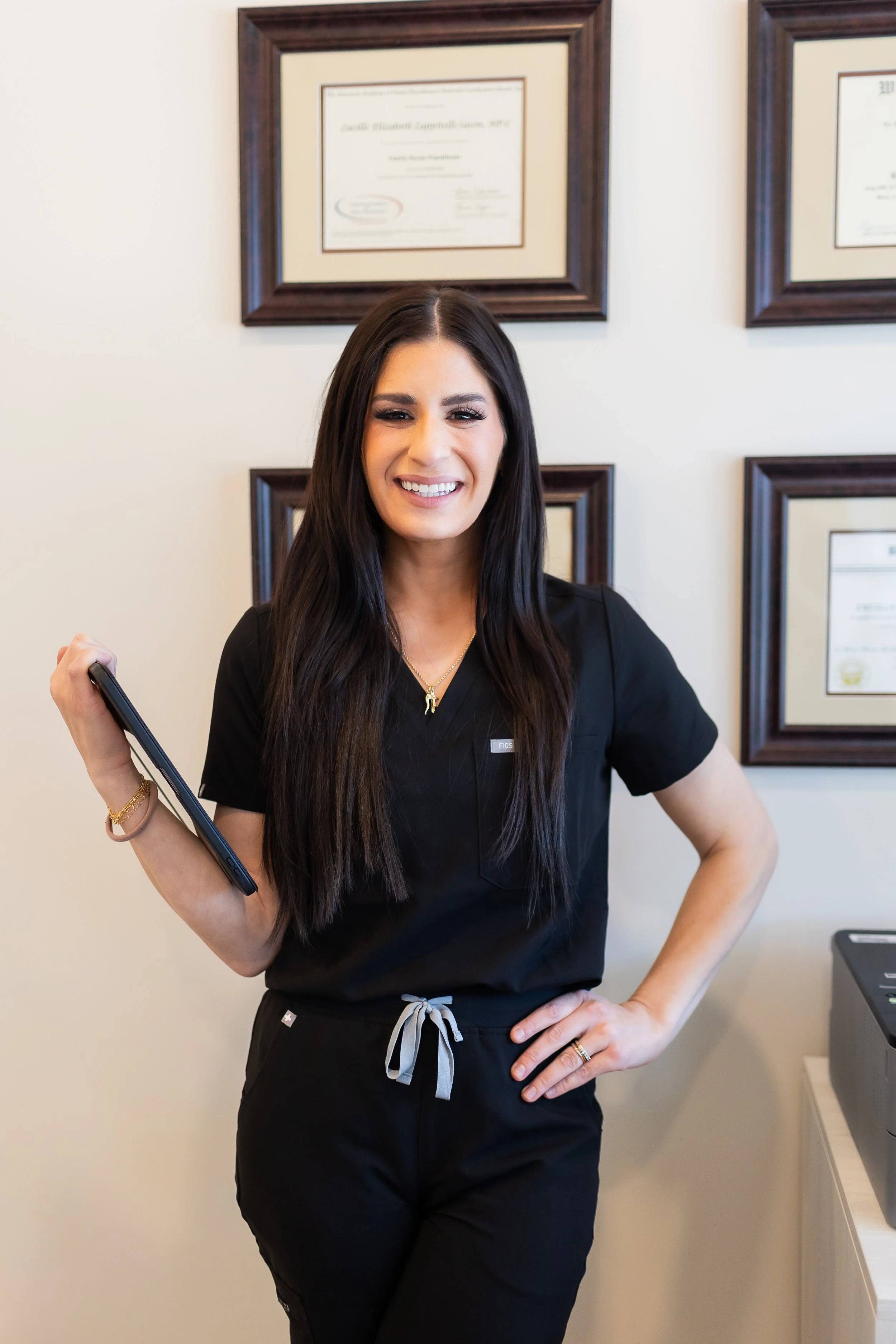 Smiling woman in black scrubs holding a tablet, standing in front of framed certificates on the wall.