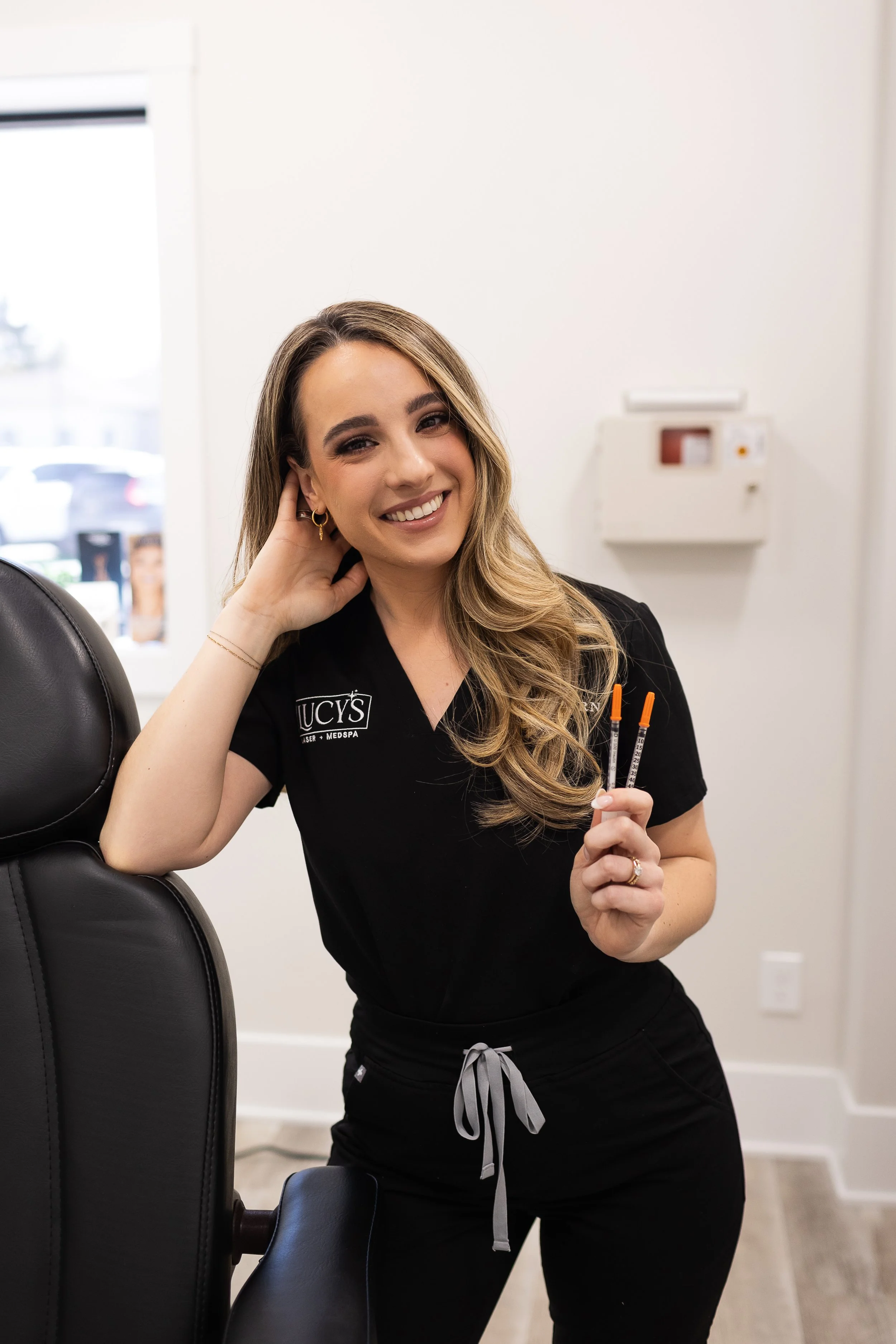 Smiling woman in black scrubs holding syringes in a medical office.