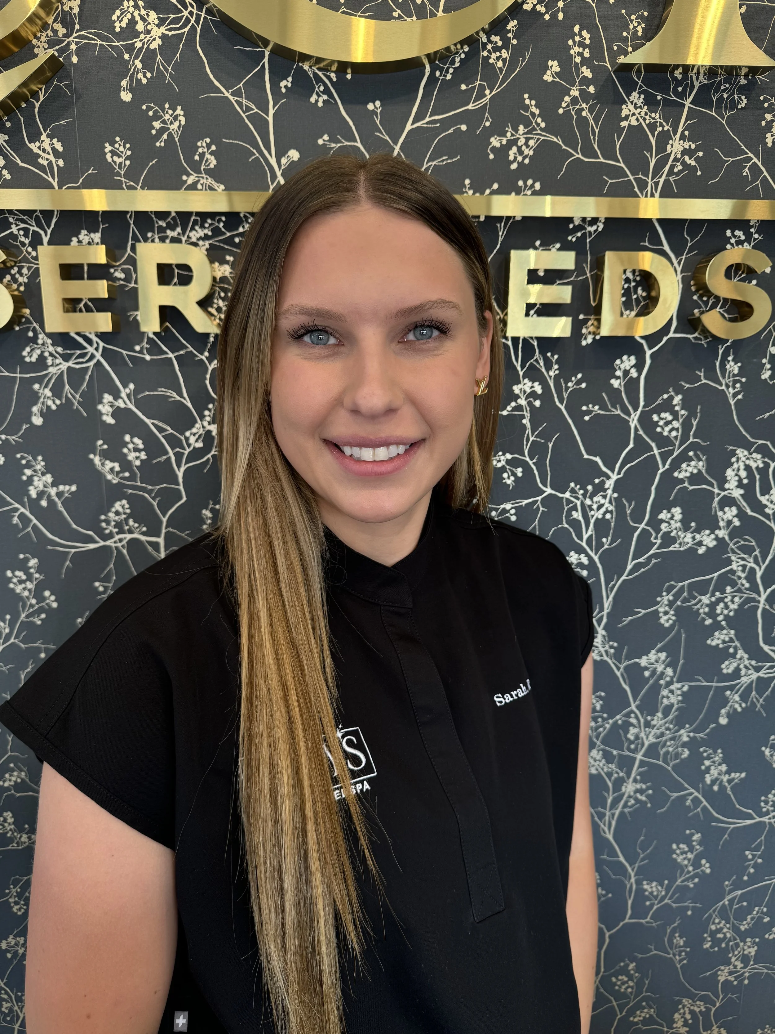 Smiling woman with long blonde hair wearing a black uniform, standing in front of a decorative wall with gold lettering and a floral pattern.