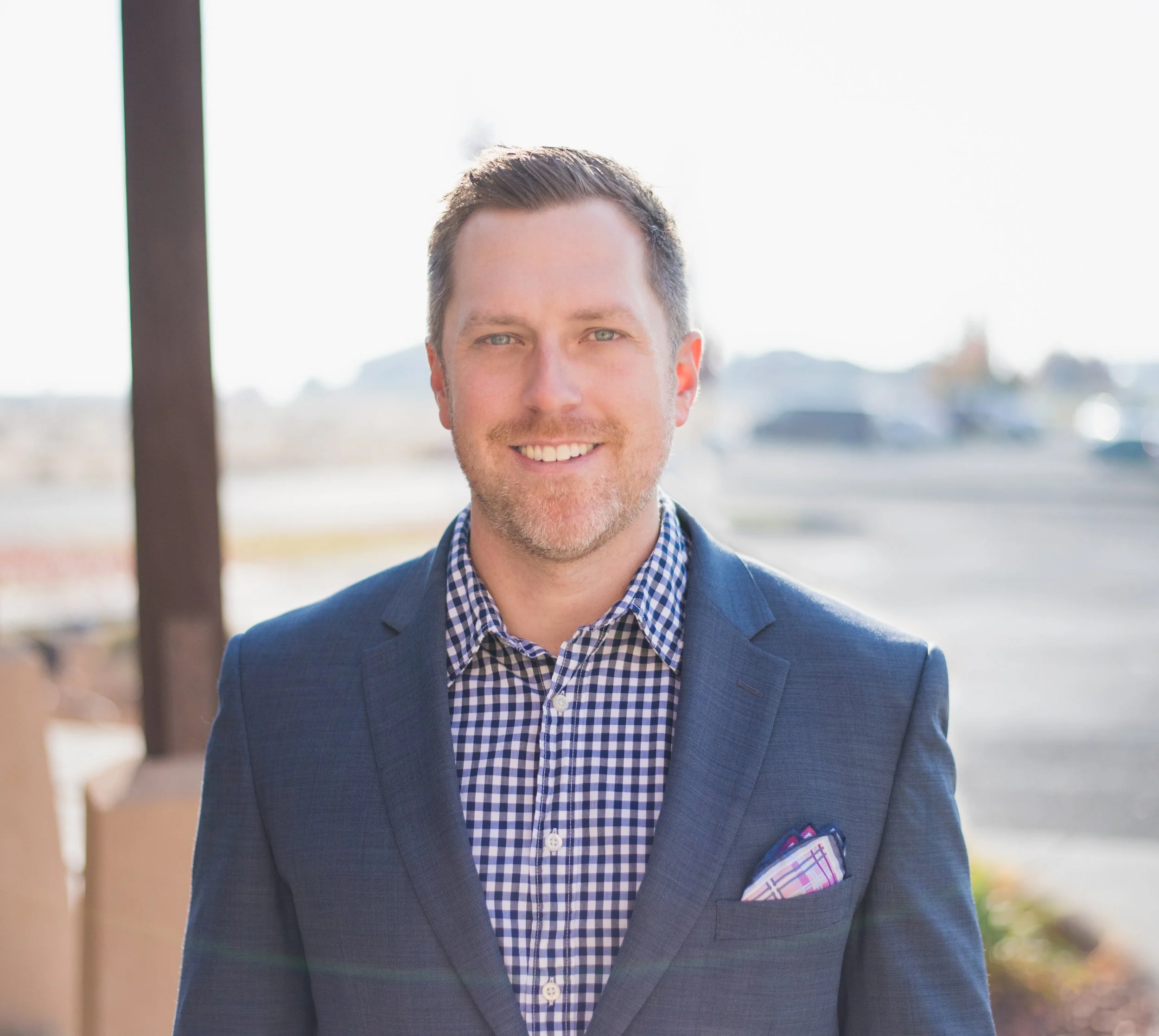 Man in blue checkered shirt and blazer smiling outdoors
