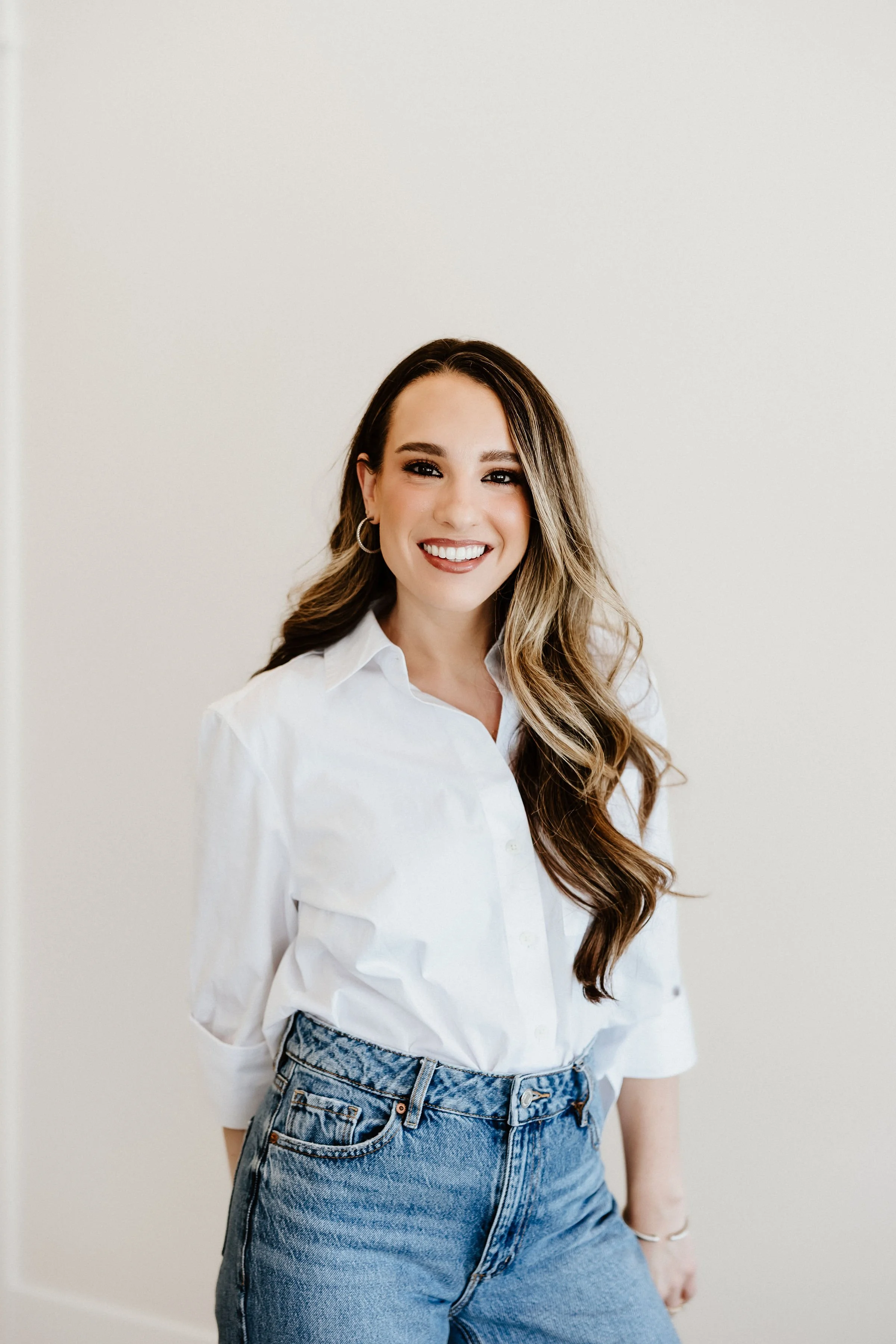A young woman with long wavy hair smiling, wearing hoop earrings, a white button-up shirt, and high-waisted blue jeans, standing against a plain white wall.