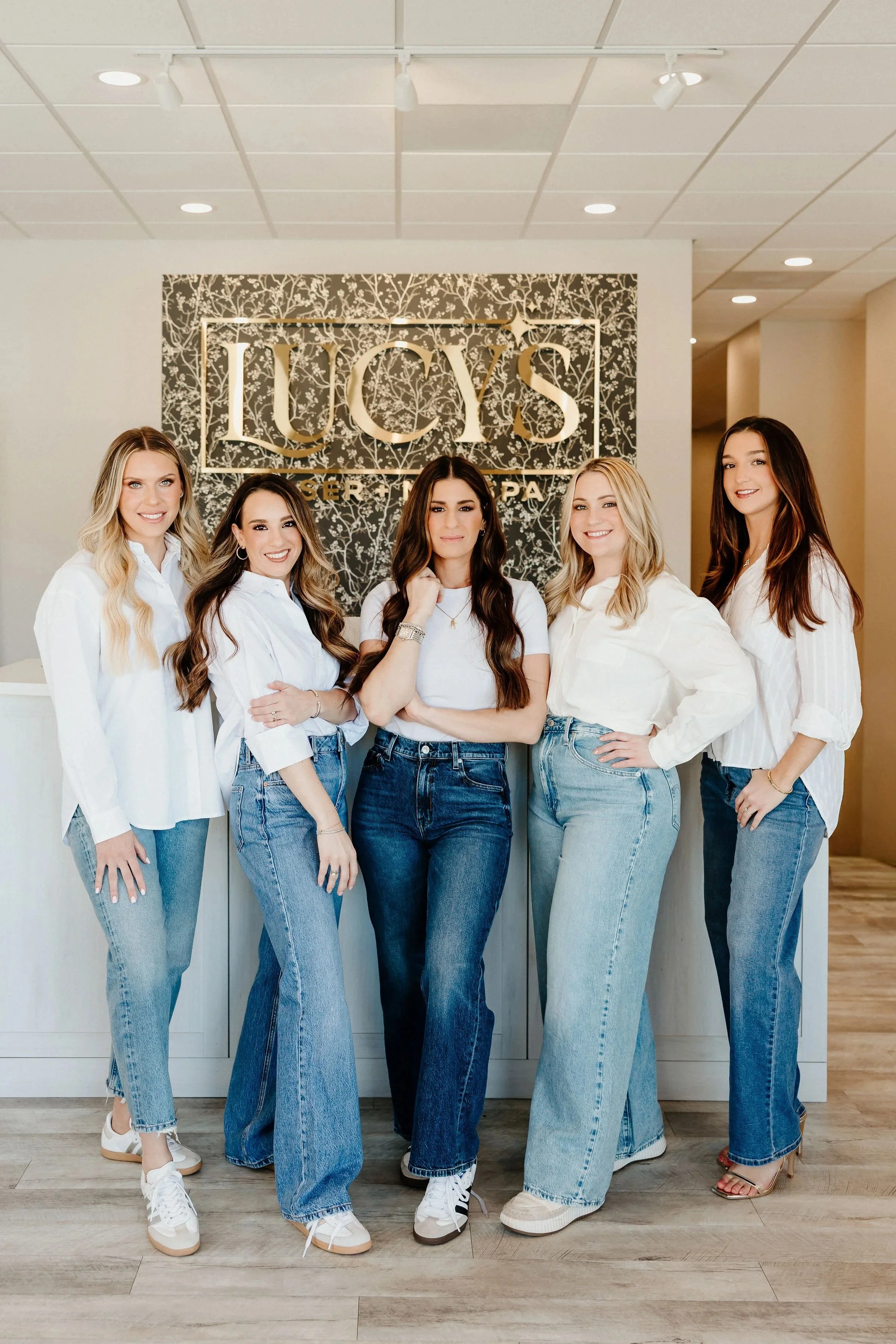 Five women standing in front of a decorative wall with the Lucy's Laser & MedSpa logo, wearing jeans and white tops, smiling for the photo.