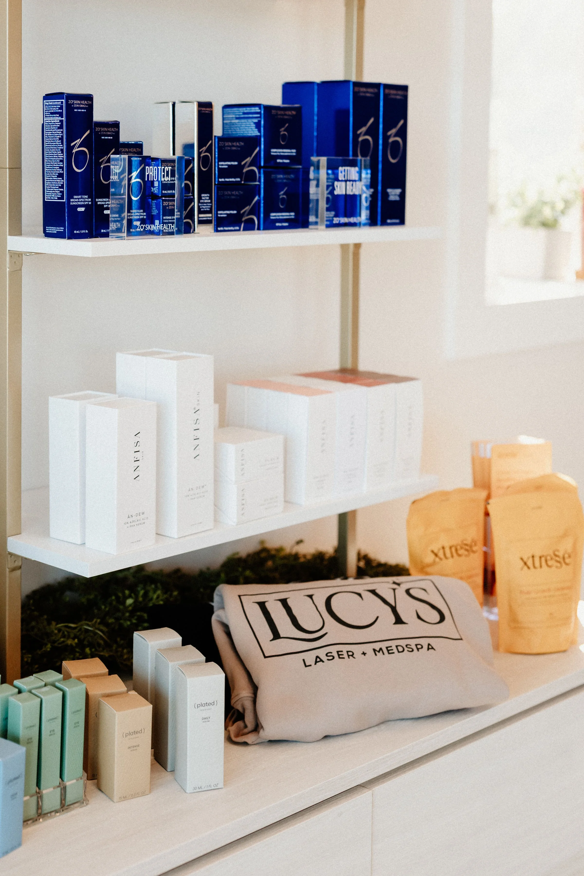 Various skincare and beauty products displayed on shelves at a store, including blue boxes and white bottles, with some promotional packaging and a cloth with the words 'LUCY'S LASER + MEDSPA' printed on it.