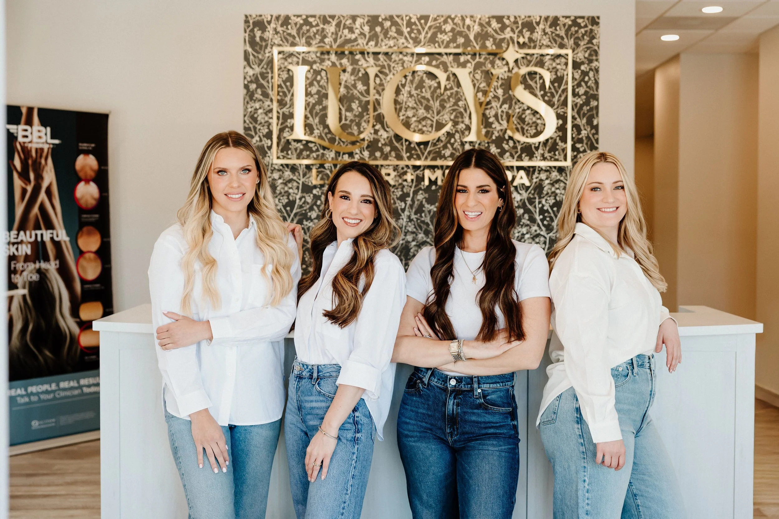 Four women standing together smiling inside the med spa, with the gold Lucy's Laser & MedSpa sign behind them.