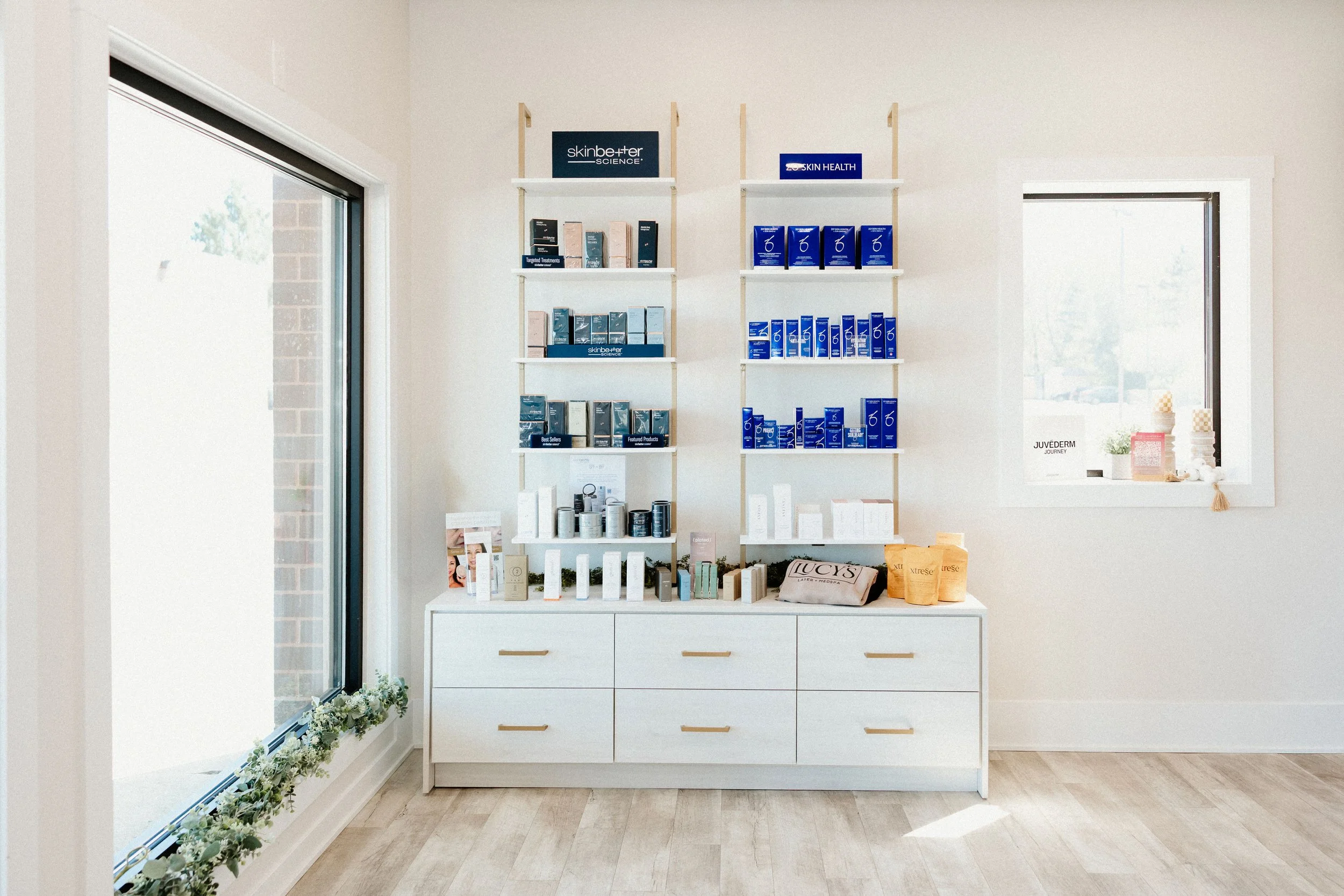 Shelves with skincare products in a minimalist salon or store, with large windows allowing natural light, beige walls, and light wood flooring.