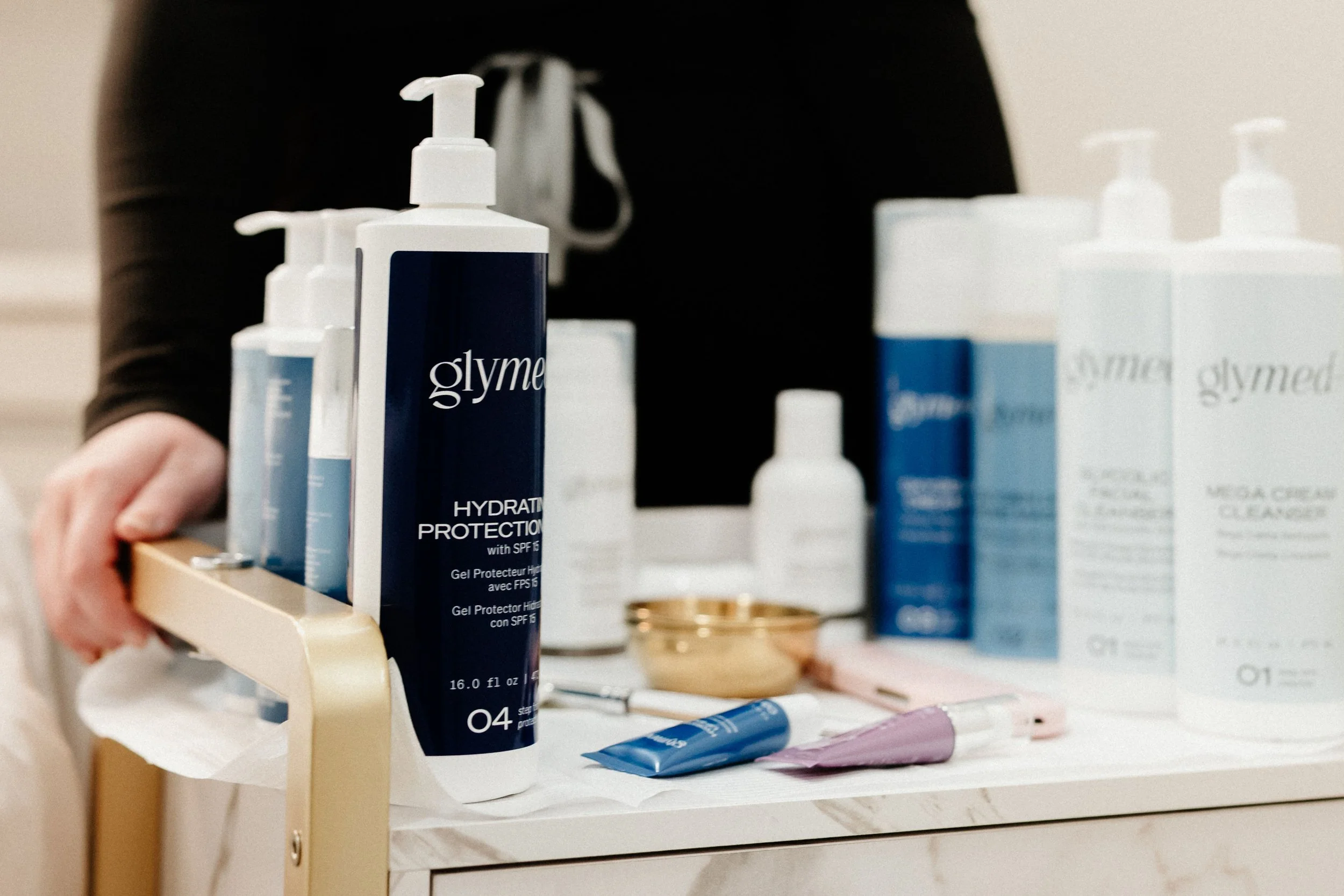 Several skincare products, including a blue bottle labeled 'glymed' Hydration Protection, are arranged on a white shelf.