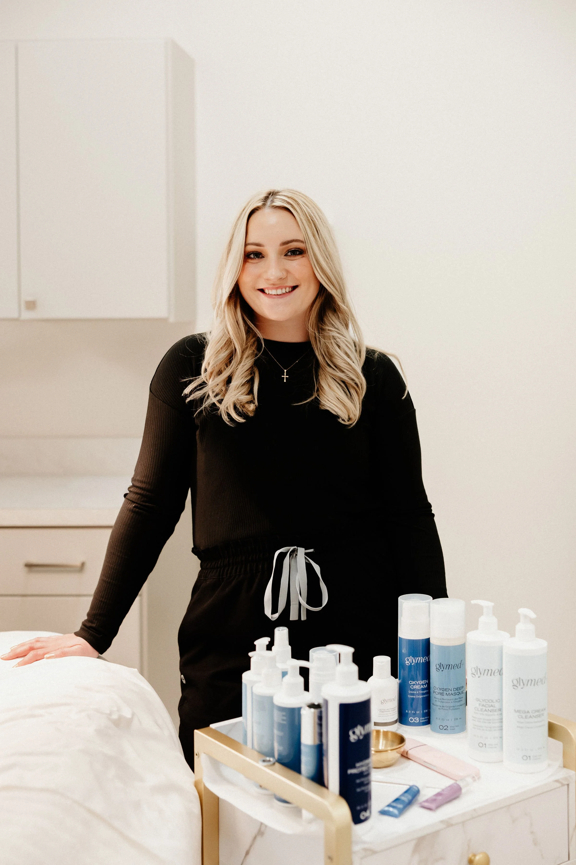 A smiling woman with blonde hair in a black long-sleeve top and cross necklace standing behind a counter with skincare products, in a clean, bright room.