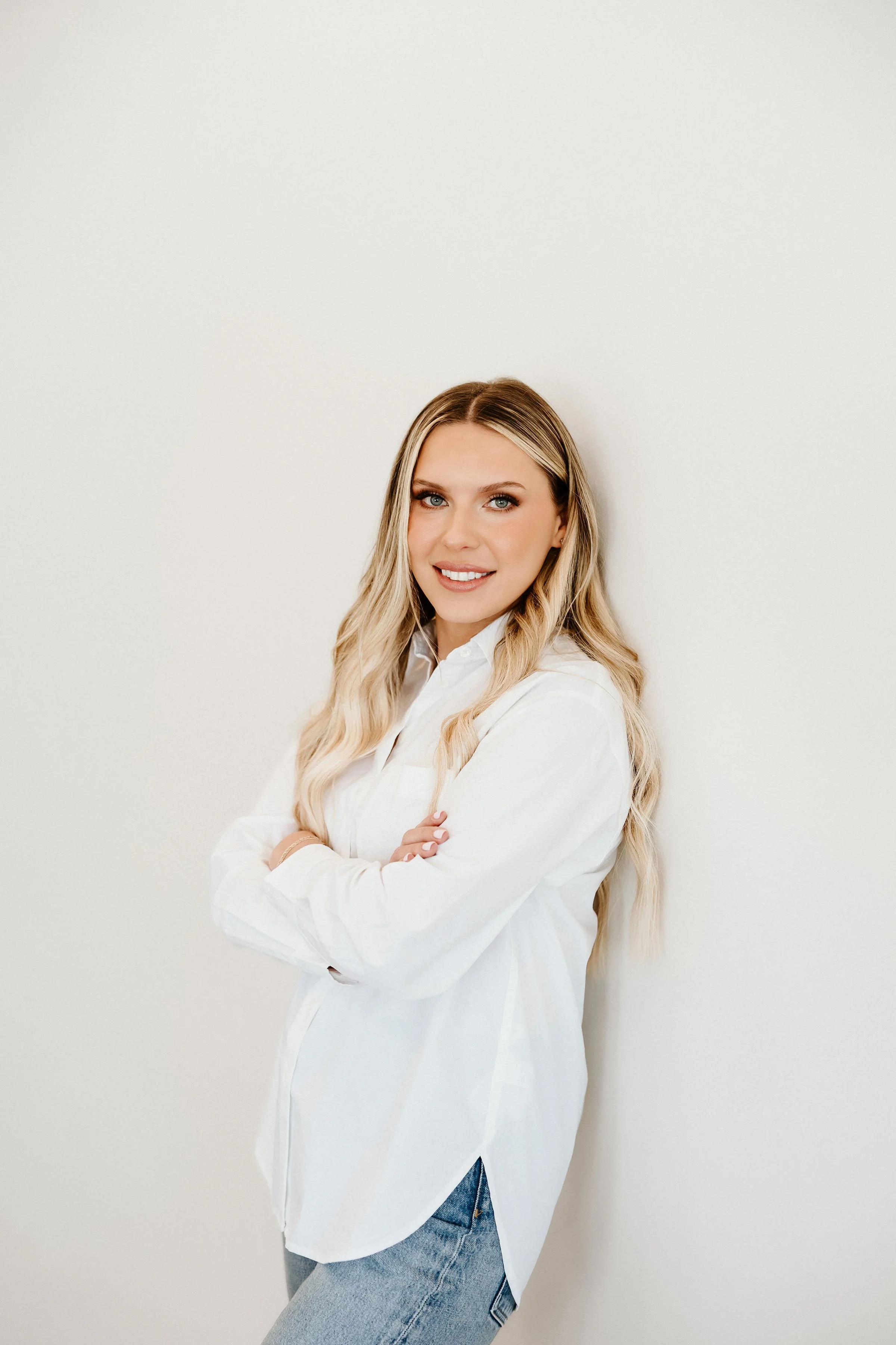 A young woman with long blonde hair, wearing a white shirt and jeans, standing cross-armed and smiling against a plain white wall.
