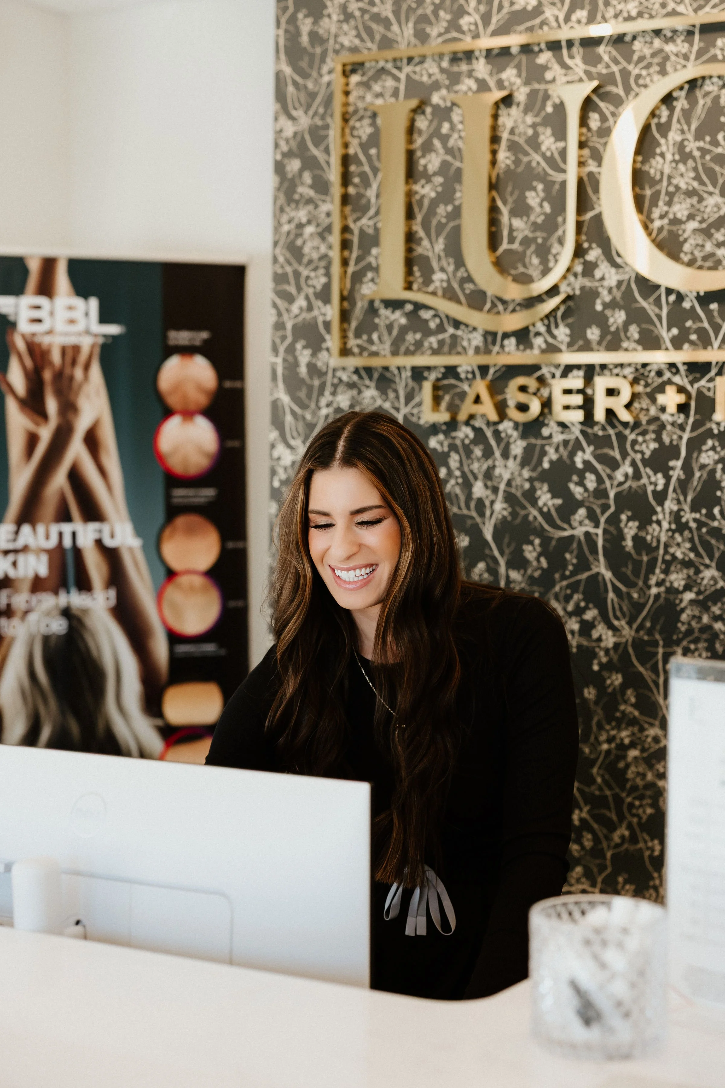 A woman with long dark hair smiling behind a white counter at a laser spa, with a large sign reading 'LUC LASER +' on a decorative floral wallpaper background.