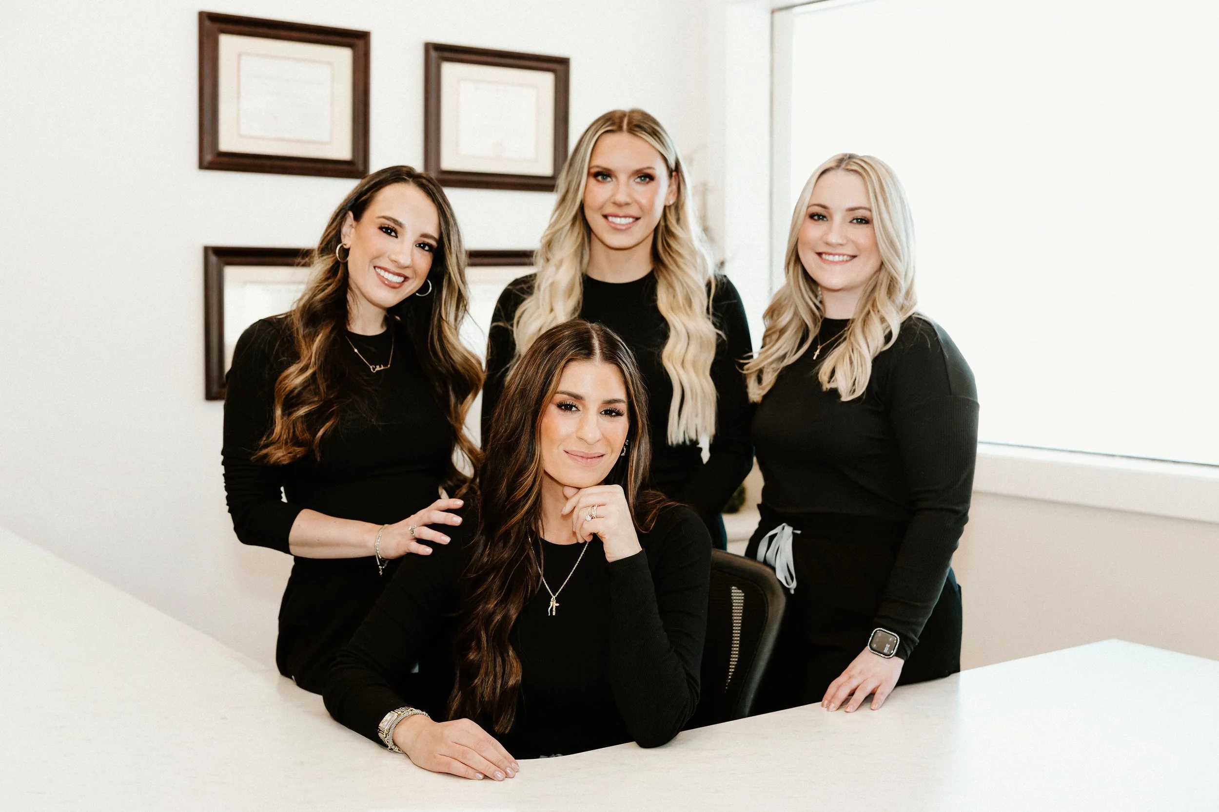 Four women with long hair wearing black shirts, posing together indoors, three standing behind a seated woman at a white table, all smiling.