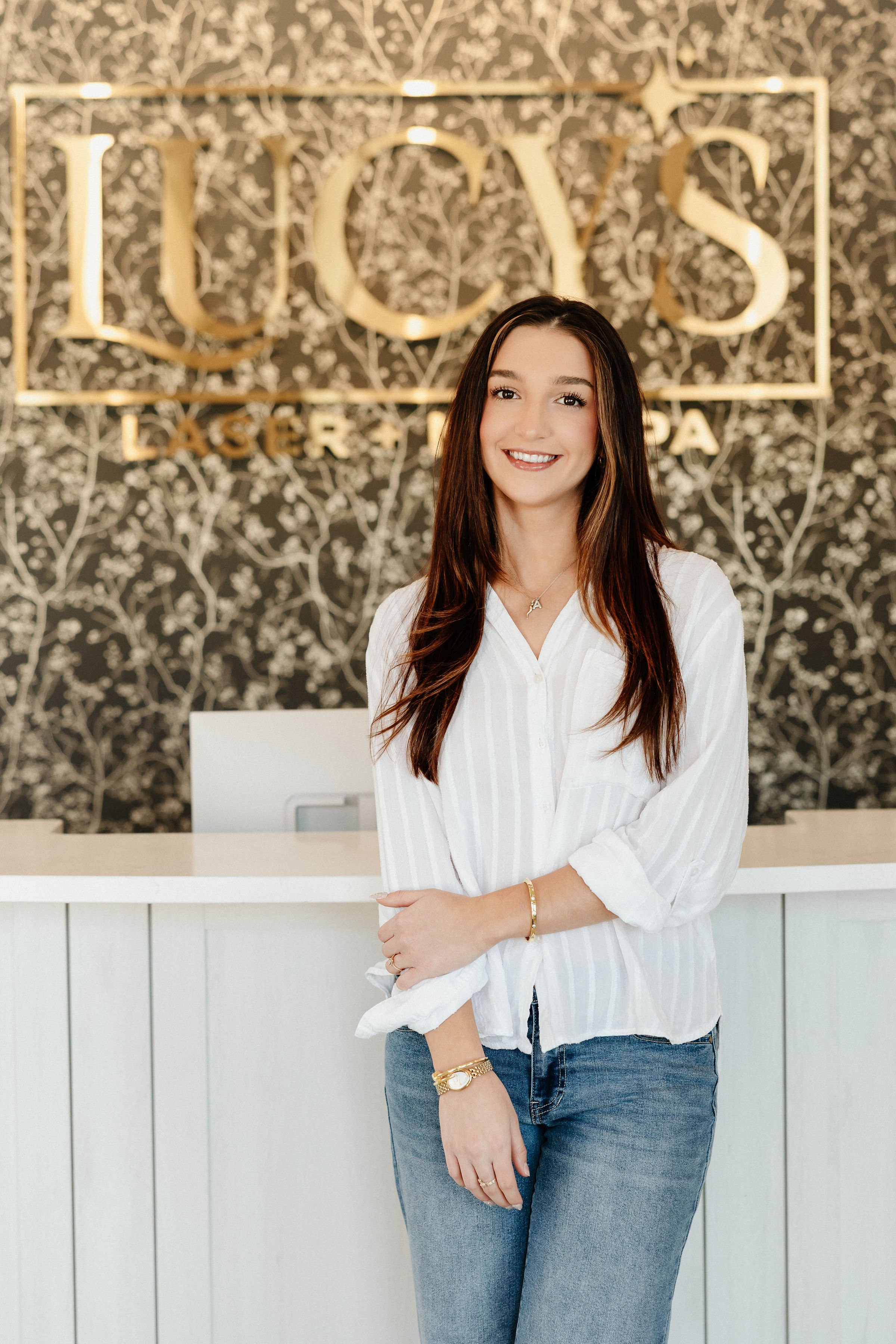 A woman standing inside a modern, stylish space with a decorative black-and-white patterned wall behind her. She has long brown hair, is smiling, and wears a white blouse, blue jeans, and jewelry, including a watch and bracelets.
