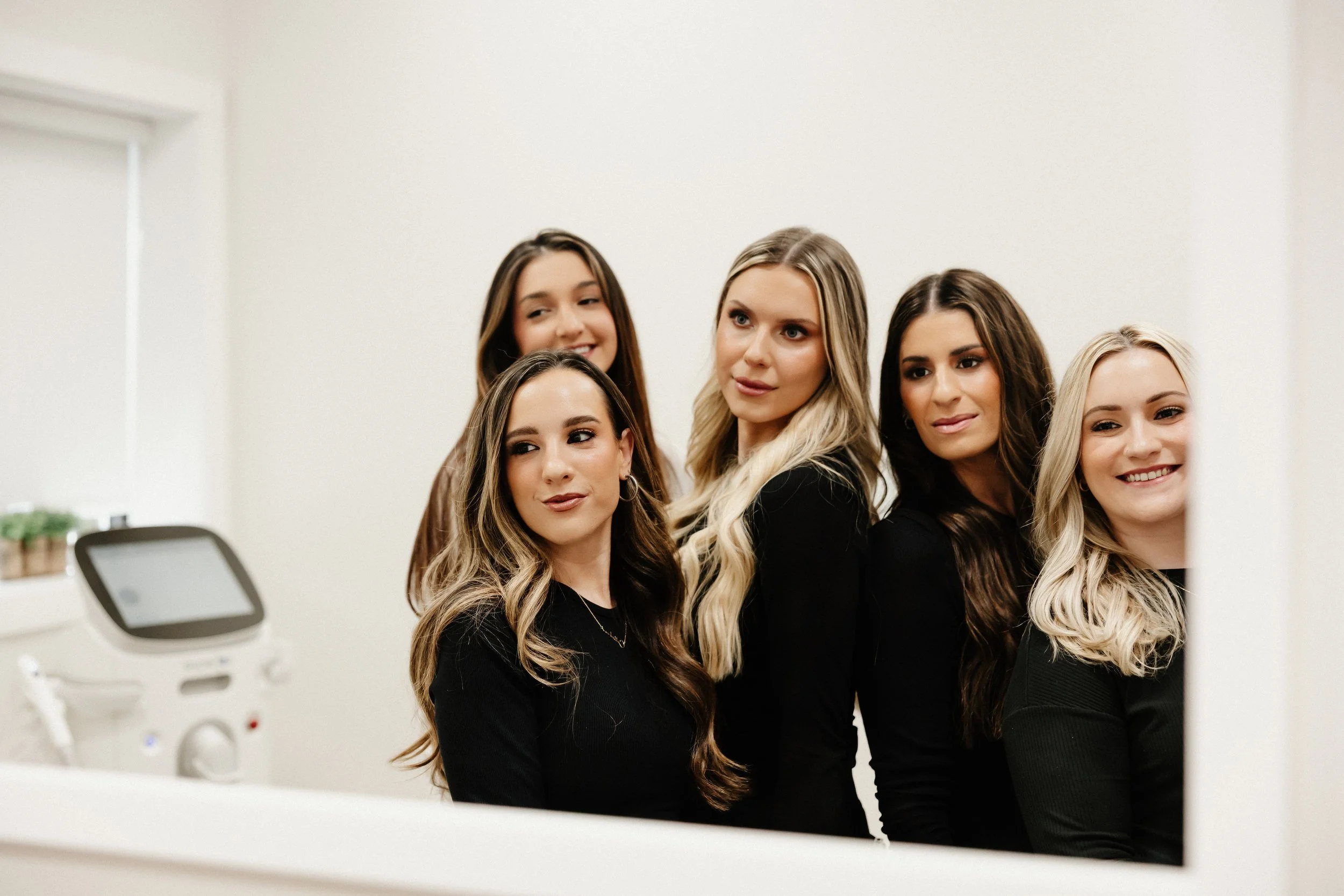 Group of five women with long, styled hair in a bright, clinical room looking at a computer screen.
