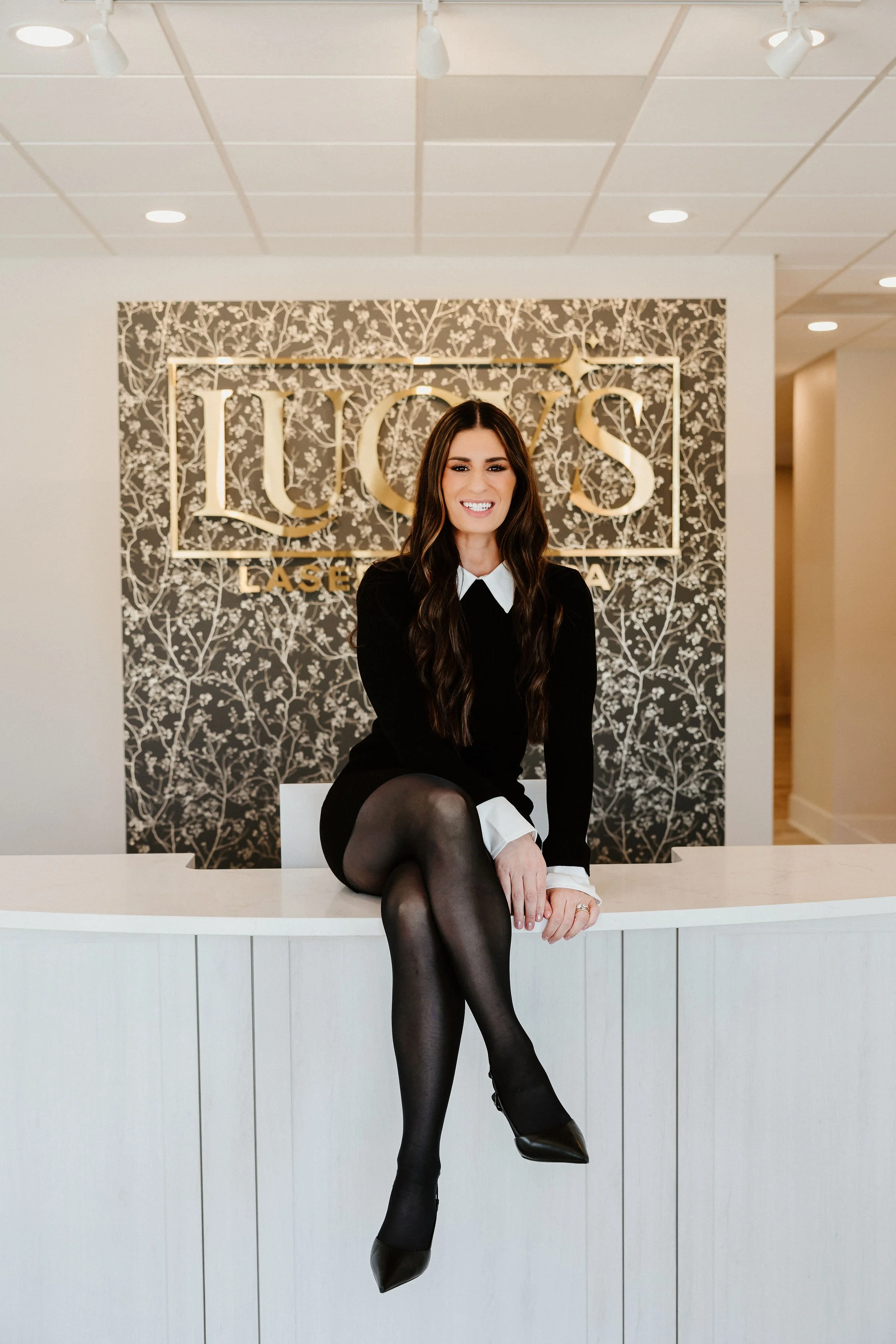 A woman with long dark hair sitting on a reception desk in front of a black and white floral patterned wall with a sign that reads 'LUCY'S LASER' in gold letters.