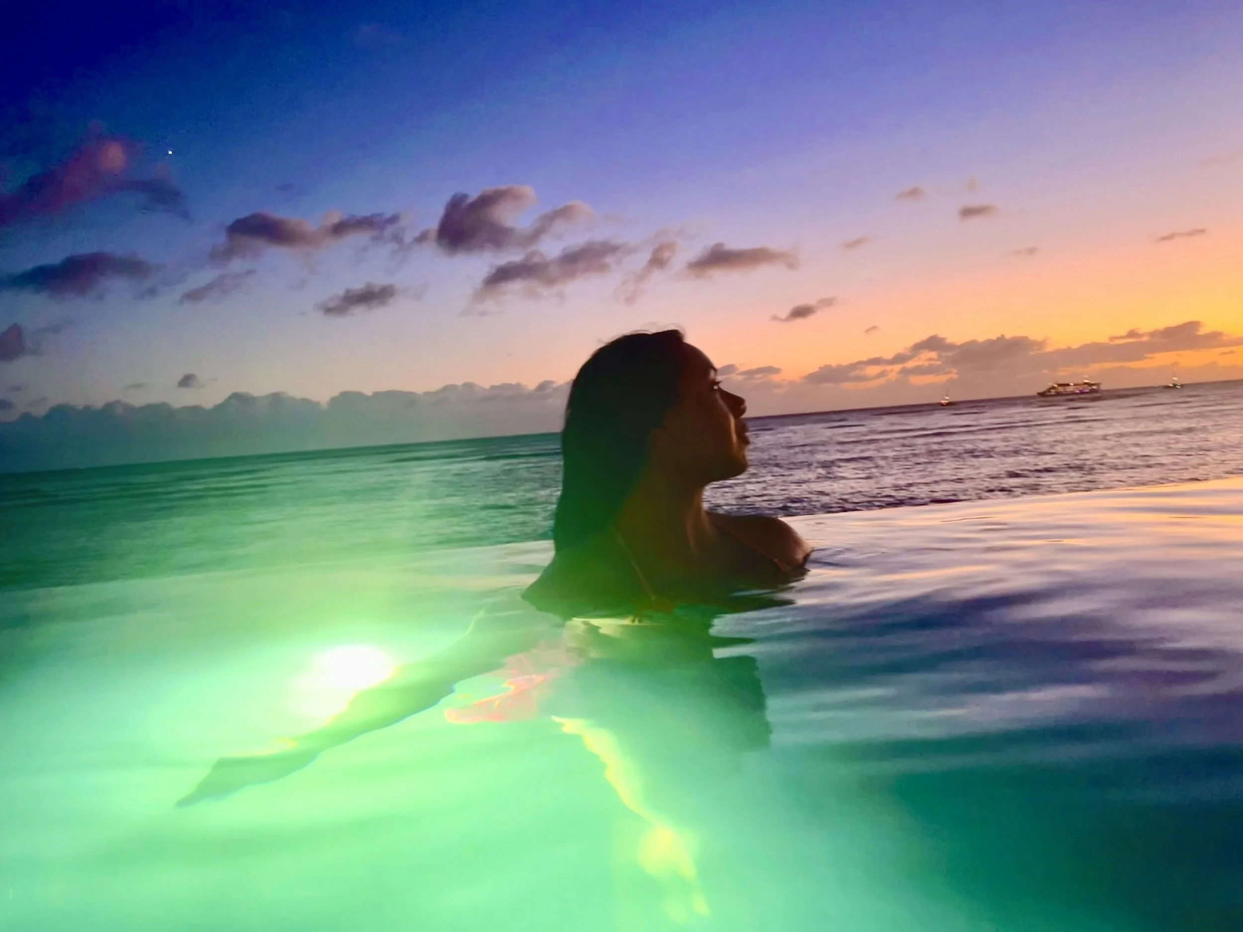 A person swimming in an infinity pool at sunset with the ocean and boats in the background.