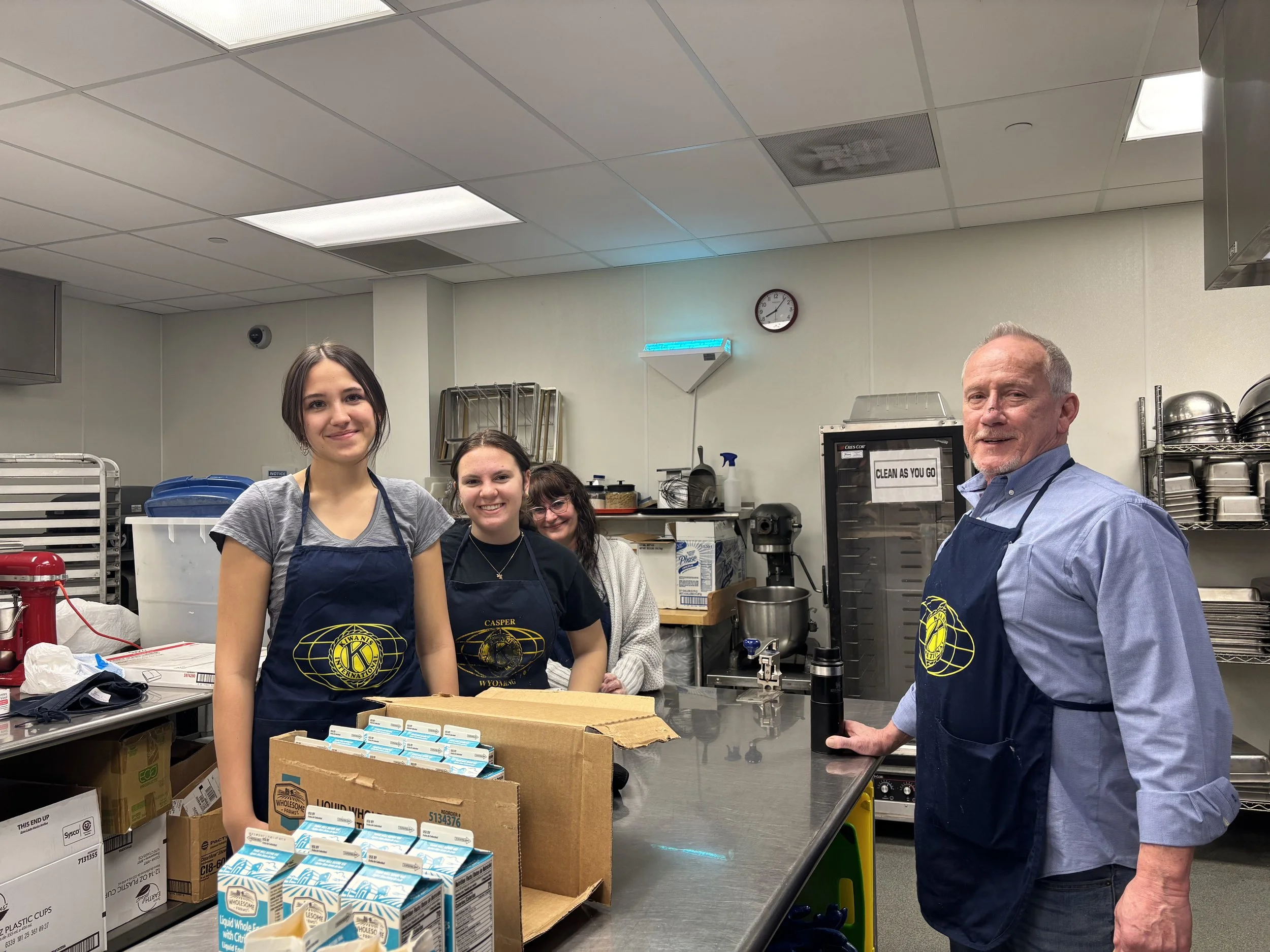 Three woman and one  man wearing Kiwanis aprons stand in an industrial kitchen.