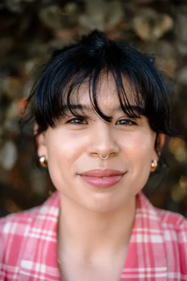 Close-up portrait of a woman with short black hair, light skin, and a septum piercing, wearing small gold earrings and a pink checkered shirt, outdoors with blurred brown and green background.