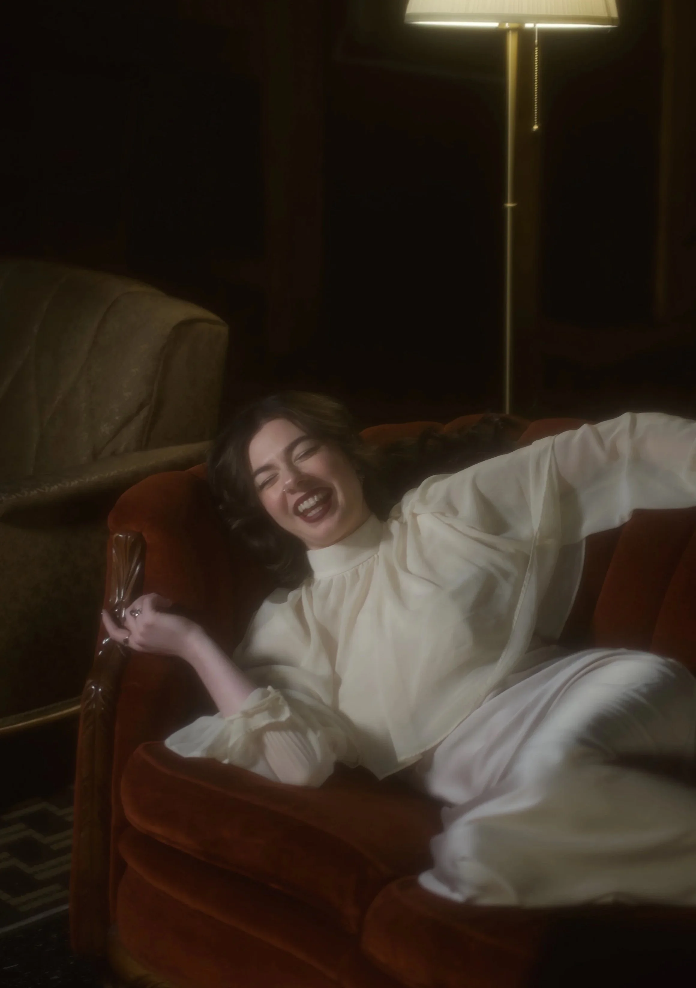 woman in white dress laughing, laying down on vintage red couch