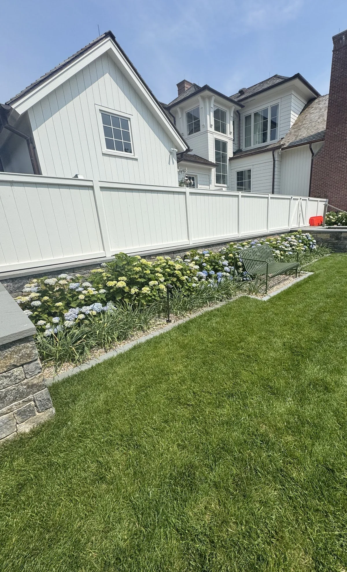 A backyard with well-maintained green grass, a flower bed with white and blue hydrangeas, a green metal bench, a white privacy fence, and large white house with multiple windows in the background under a blue sky.