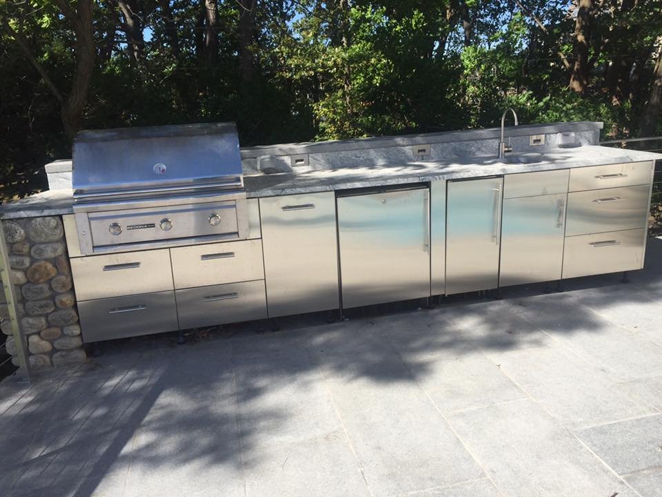 Outdoor stainless steel kitchen counter with grill, sink, and storage cabinets, set against a background of green trees.